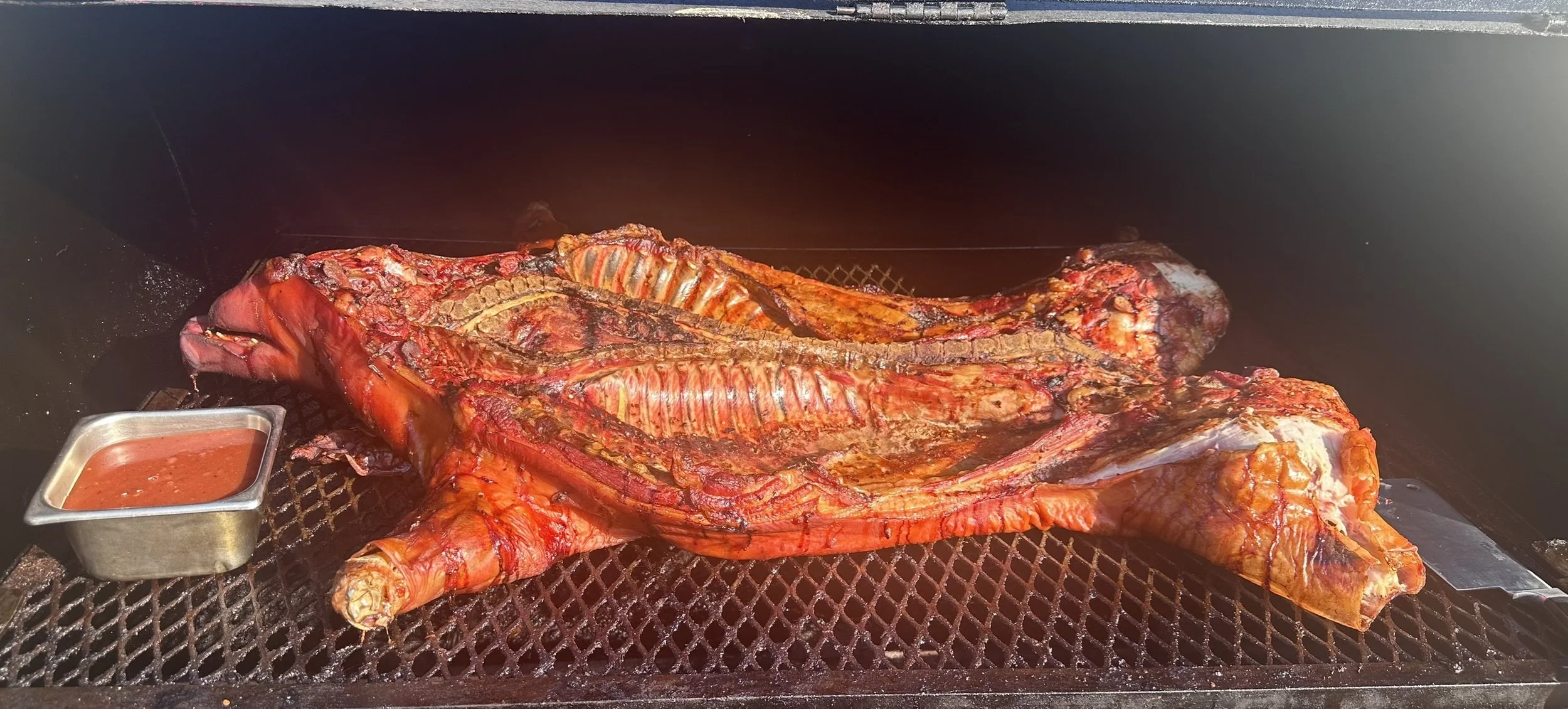 Whole large cut of beef being smoked or cooked in an outdoor grill or smoker, with a small container of barbecue sauce beside it.