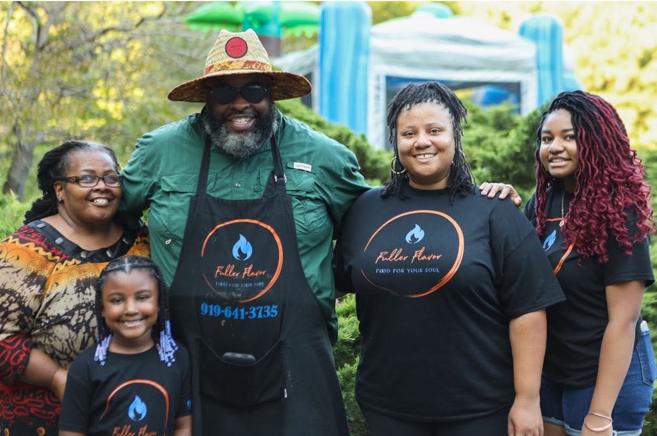 Group of five people, including two children, posing outdoors at a park or festival. They are smiling, with three women, one man, and a young girl. The man is wearing a straw hat, sunglasses, a green shirt, and a black apron with a logo and text. The