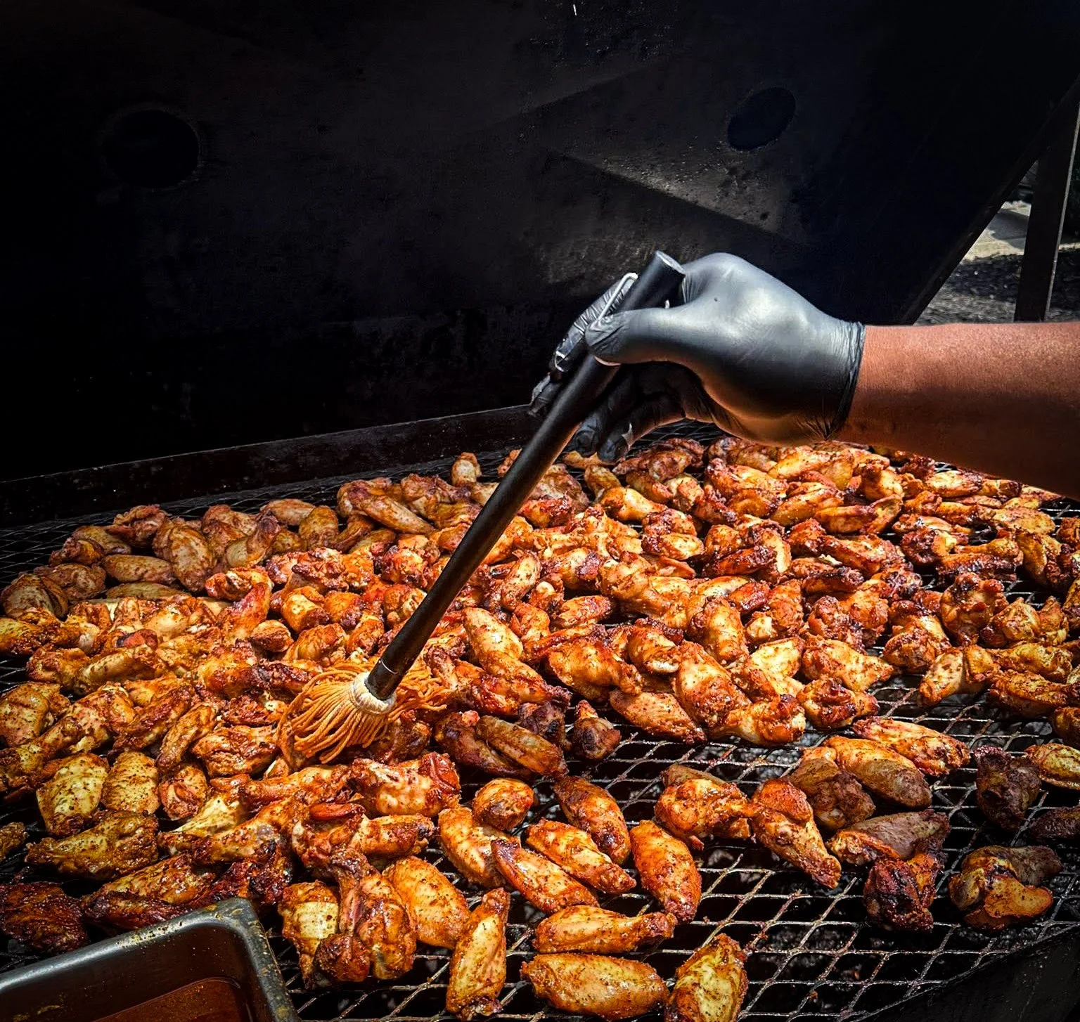 Person wearing black gloves using a brush to baste grilled chicken wings on a barbecue grill.