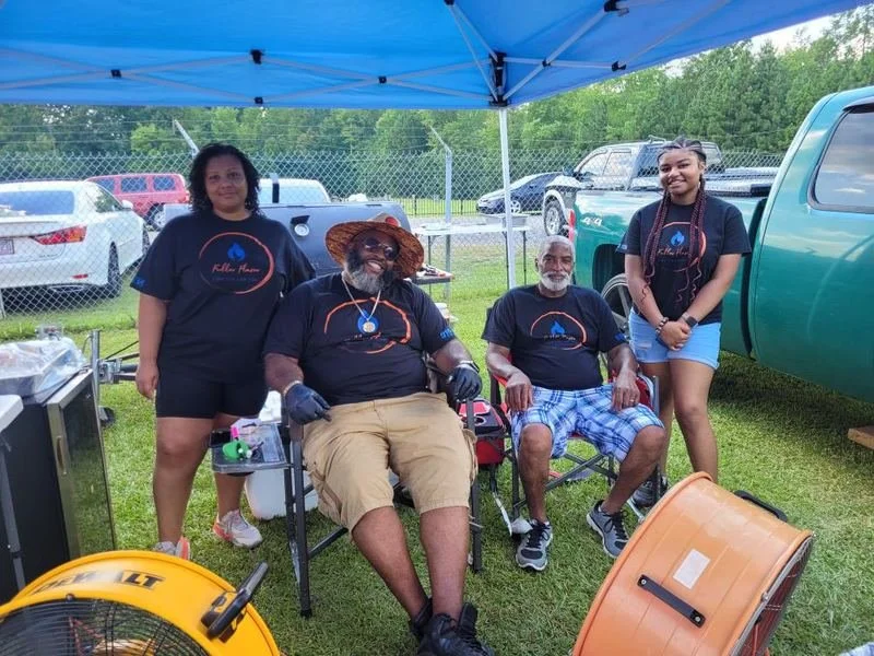 Group of four people, two men sitting and two women standing, under a blue canopy at an outdoor event with parked cars and a grassy area in the background.