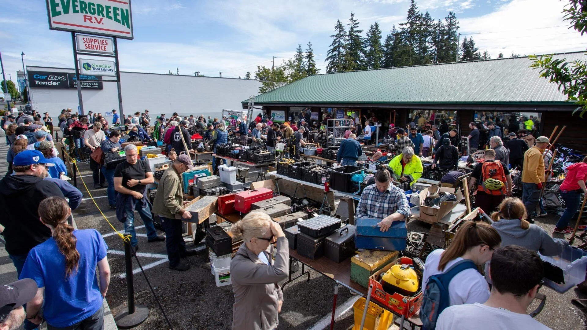 Crowd of people shopping at an outdoor garage sale or flea market with tables filled with various items, in front of a store named Evergreen R.V. with trees in the background.