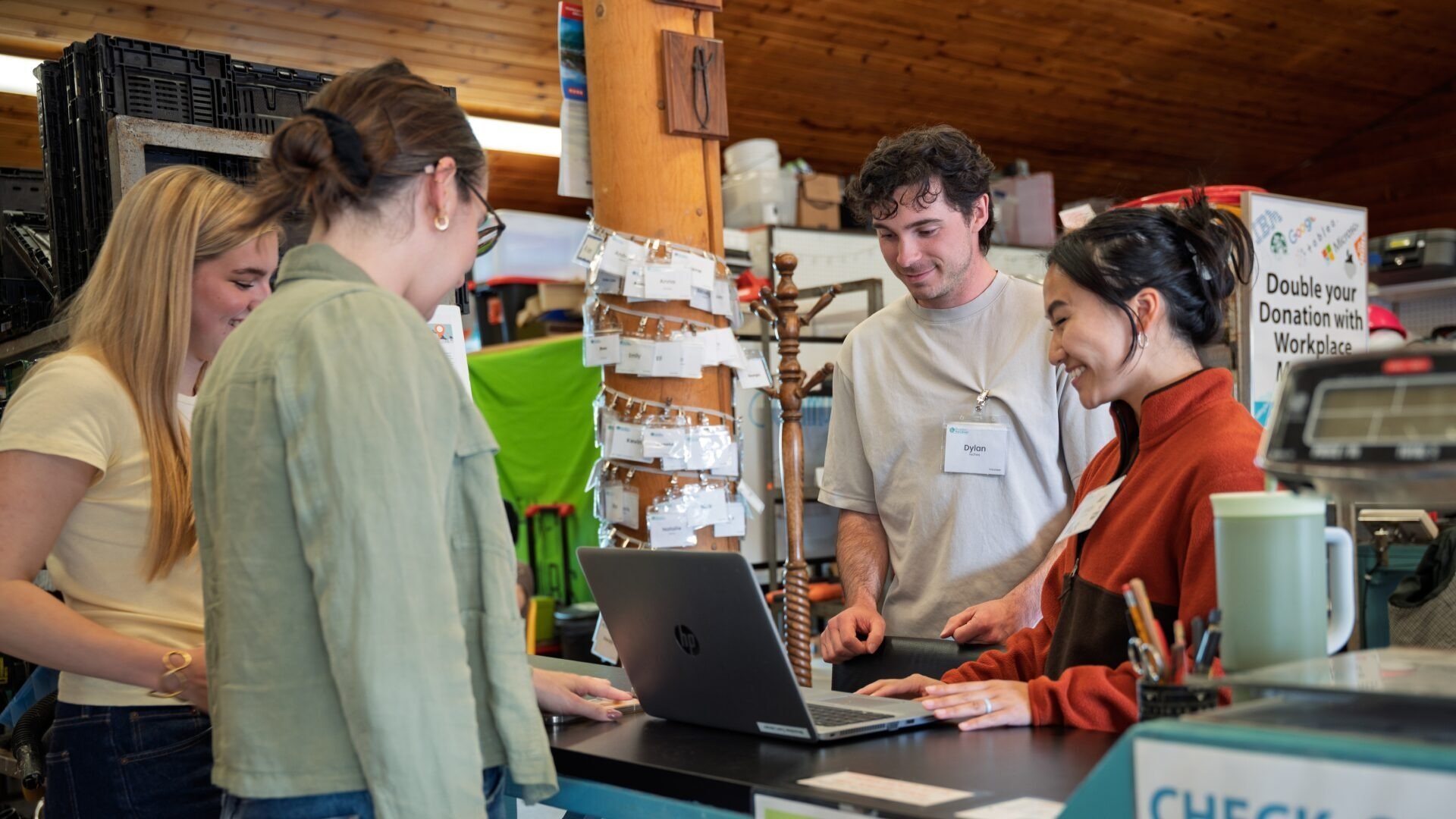 A group of people gathered around a counters at a store or office, with one person working on a laptop. They are smiling and engaging with each other. There are various supplies, signs, and equipment around them.