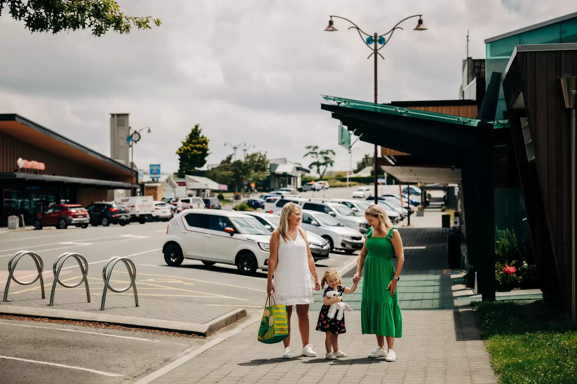 Two women and a young girl walking on a sidewalk in a shopping area with cars parked in the background. One woman is wearing a white dress and carrying a shopping bag, the other woman is in a green dress, and the girl is holding the woman in green's hand.