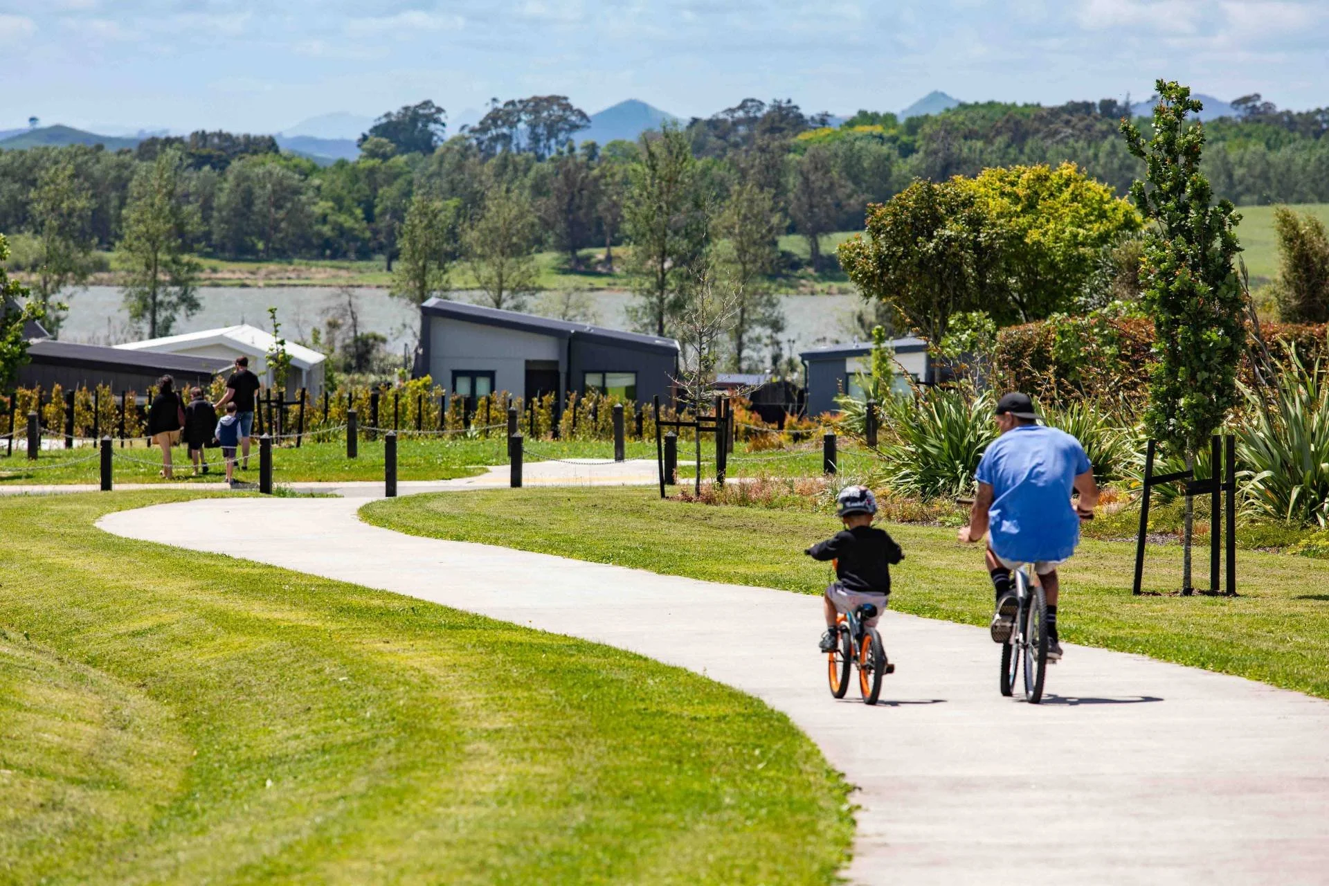 People riding bicycles and walking on a winding path in a park with trees, a lake, and distant hills in the background.