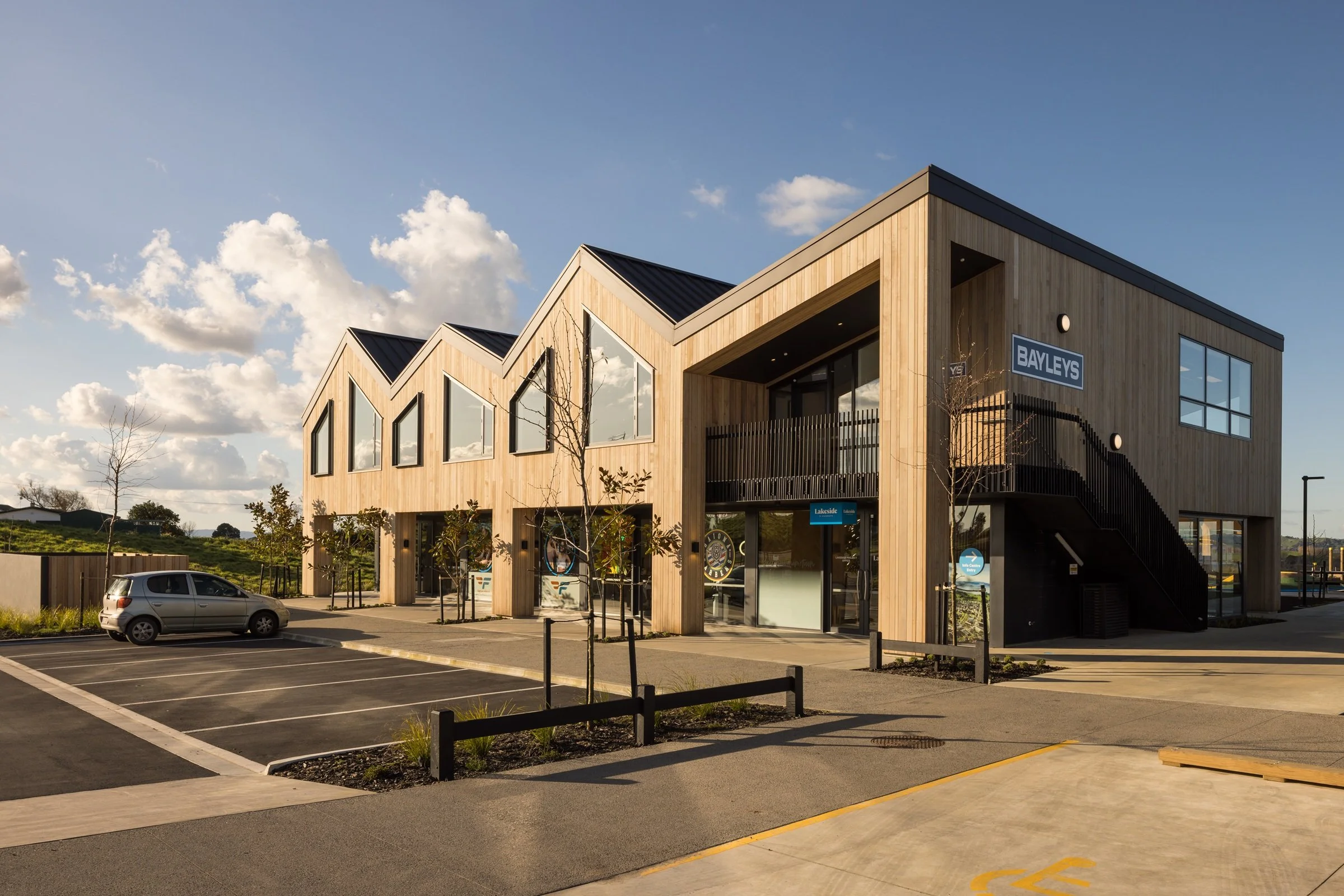 Modern building with wooden facade and large windows, a parking lot in front, and a clear sky with scattered clouds.