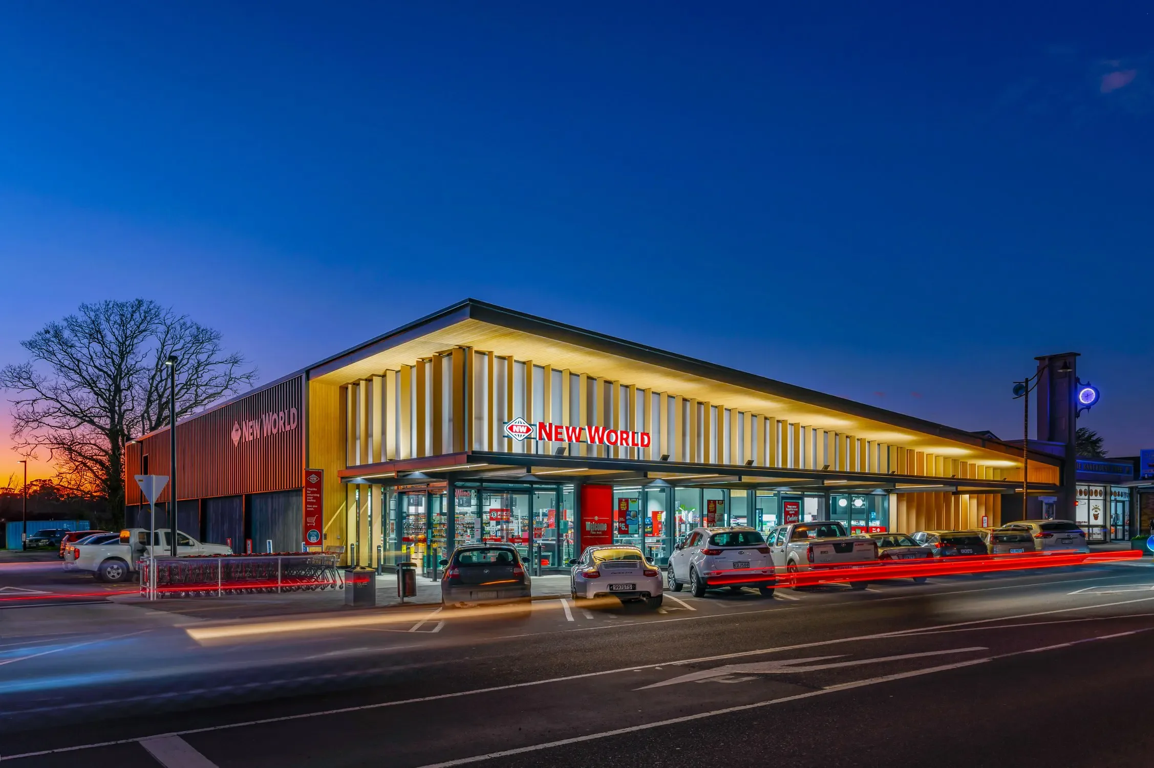 A modern grocery store named 'New World' at sunset with a parking lot filled with cars and light trails from moving vehicles.