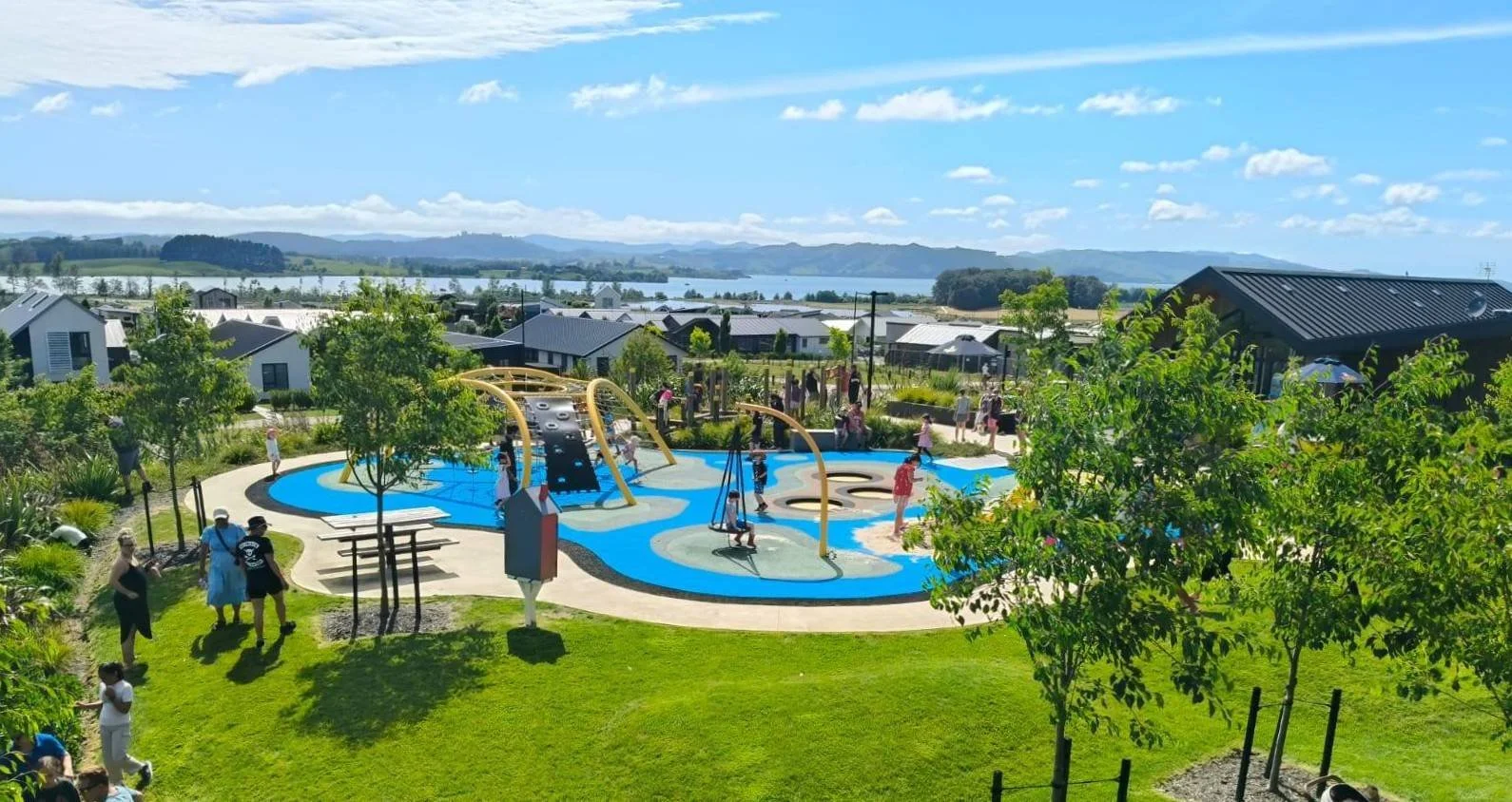 Children playing on a colorful outdoor water play area with slides, climbing structures, and swings, surrounded by trees, with a residential neighborhood and a body of water in the distance under a blue sky with scattered clouds.