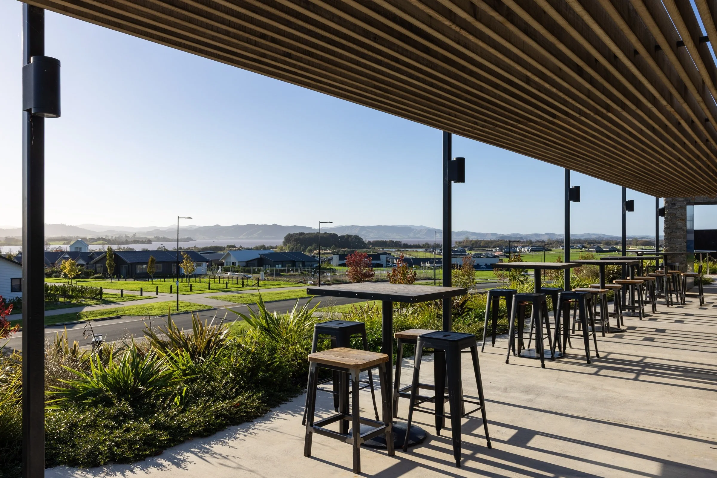 Outdoor patio with tables and barstools under a wooden pergola, overlooking a landscape with residential houses, trees, and mountains in the distance on a sunny day.