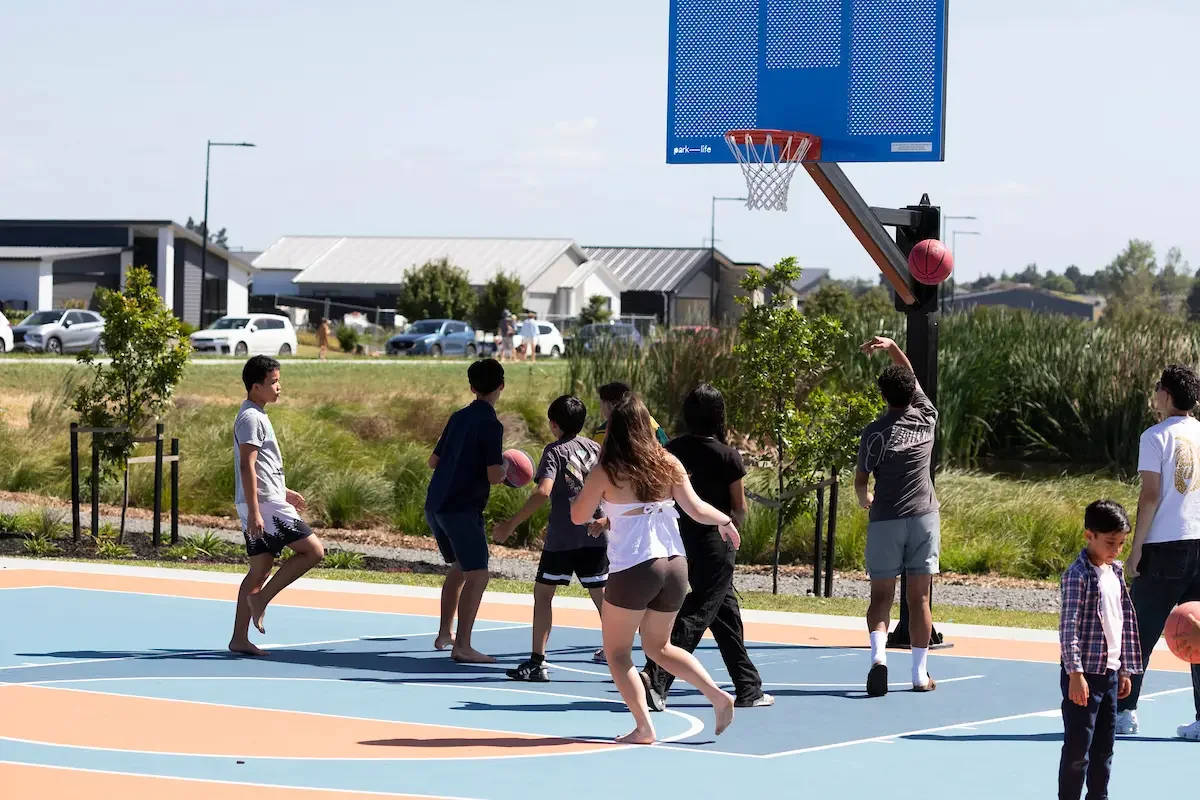 Children playing basketball on an outdoor court with a blue backboard and hoop, under a clear sky.