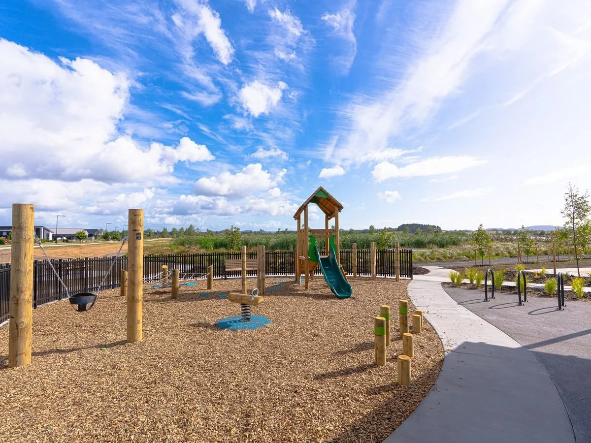 Empty playground with a slide, swings, and climbing structures, surrounded by a black fence, in a park with a winding path and open field under a partly cloudy sky.