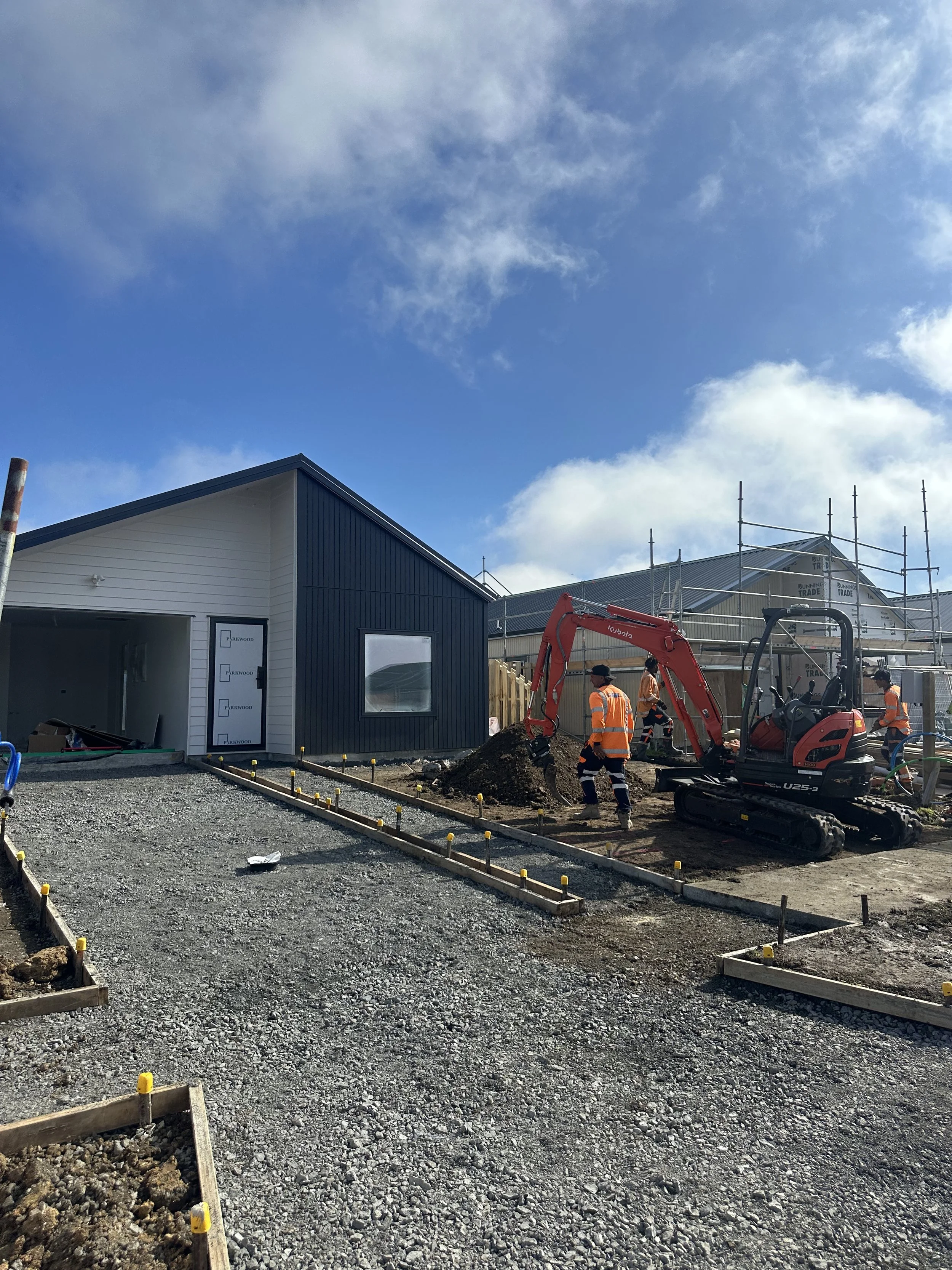 Construction site with workers and a small excavator, building frames, partially completed buildings, and a blue sky with clouds.