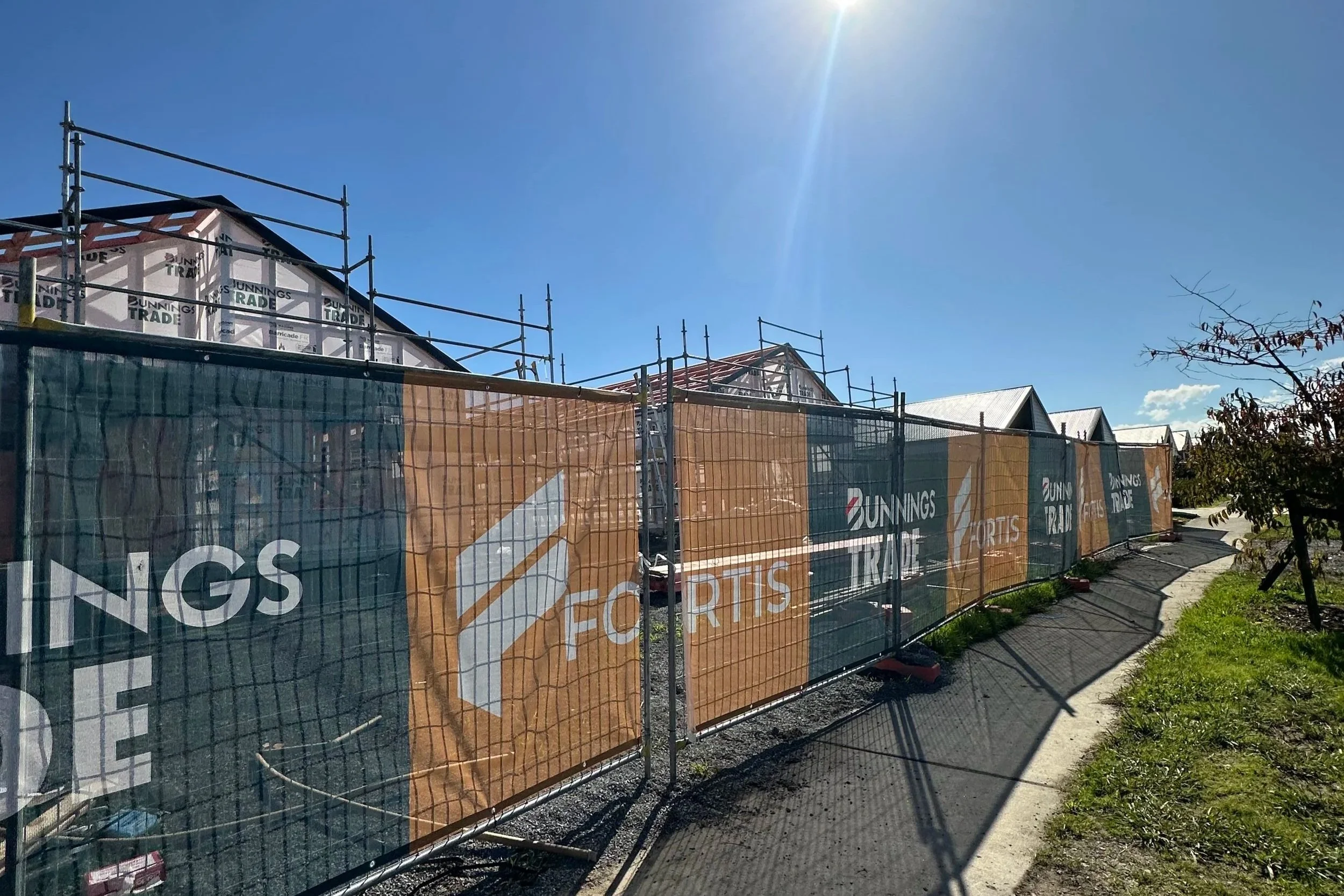 Construction site with houses in progress, surrounded by orange and green fencing with 'Bunnings Trade' branding, under a clear blue sky with the sun shining.