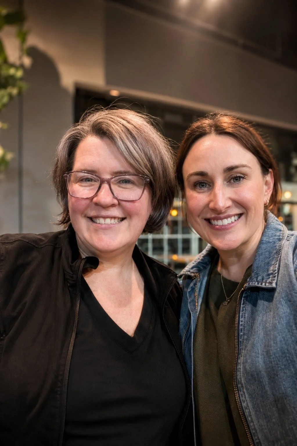 Two smiling women standing close together indoors, one with glasses and short gray hair, the other with shoulder-length brown hair, wearing a denim jacket.