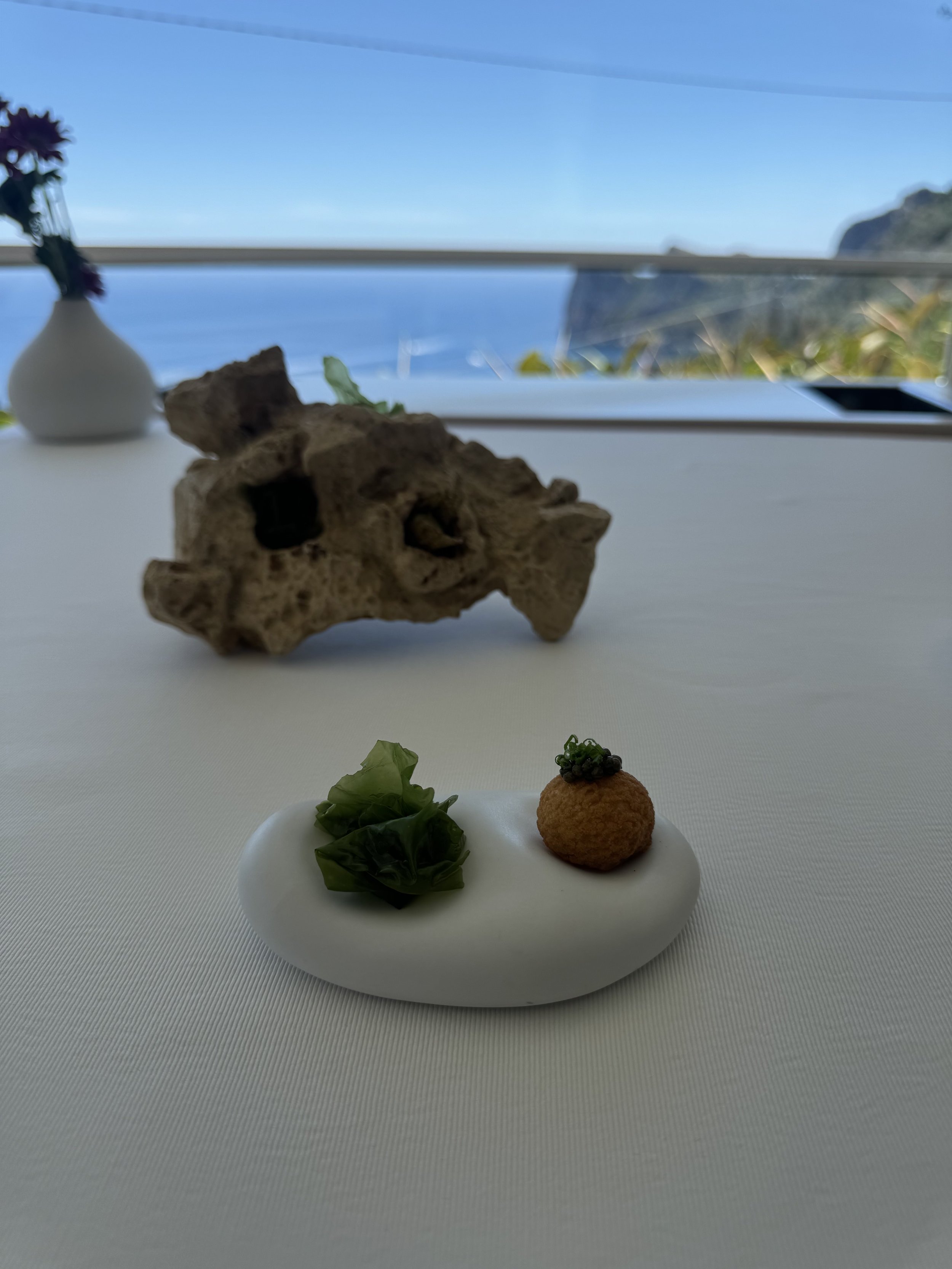 A white plate with a breaded ball topped with caviar, a small portion of greens, and a dollop of sauce, placed on a white table with a view of the ocean and a rocky coastline through a window.