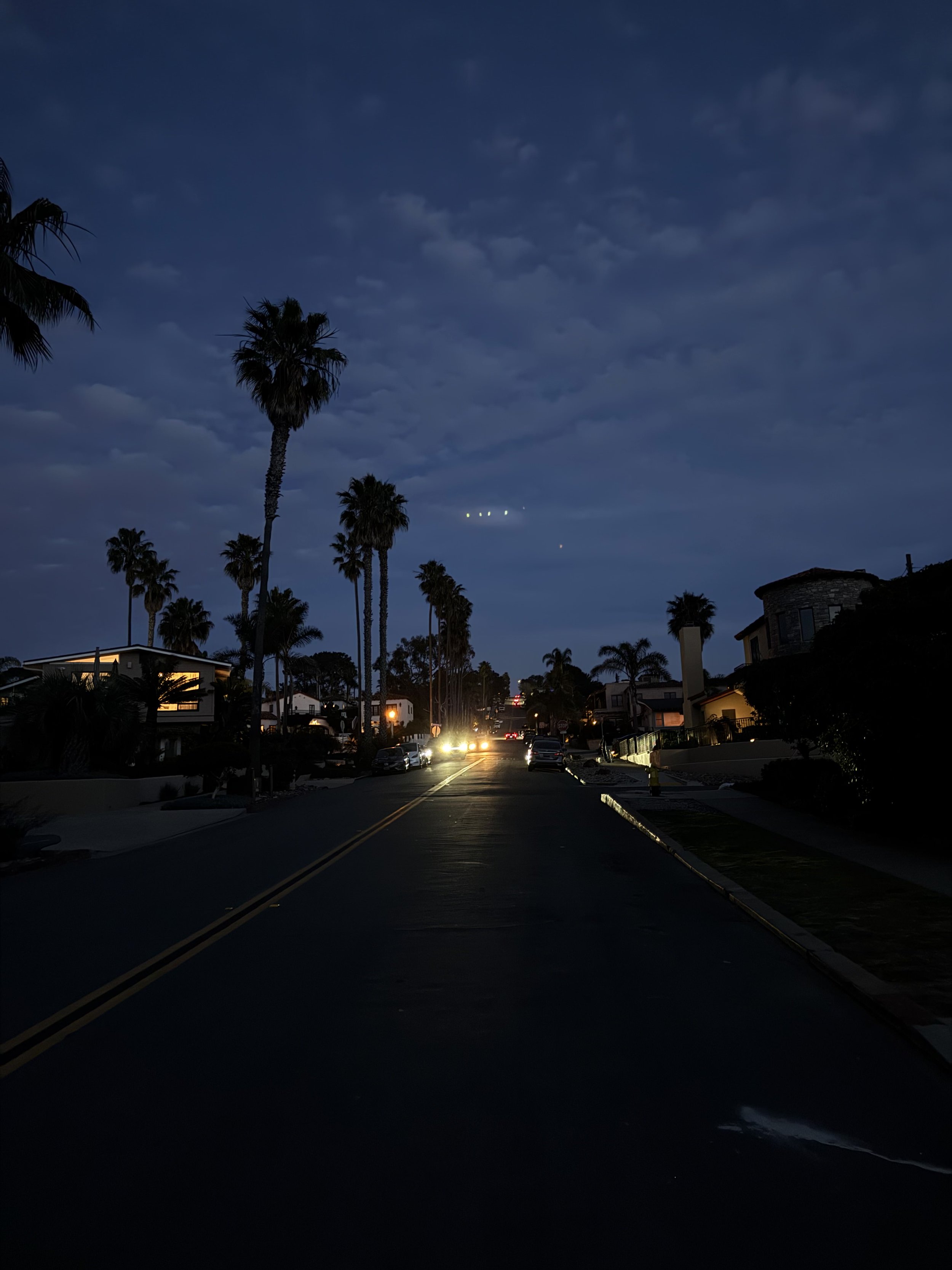 A street scene at dusk with palm trees lining the road, houses on either side, and cars with headlights on. The sky is dark with some clouds visible.