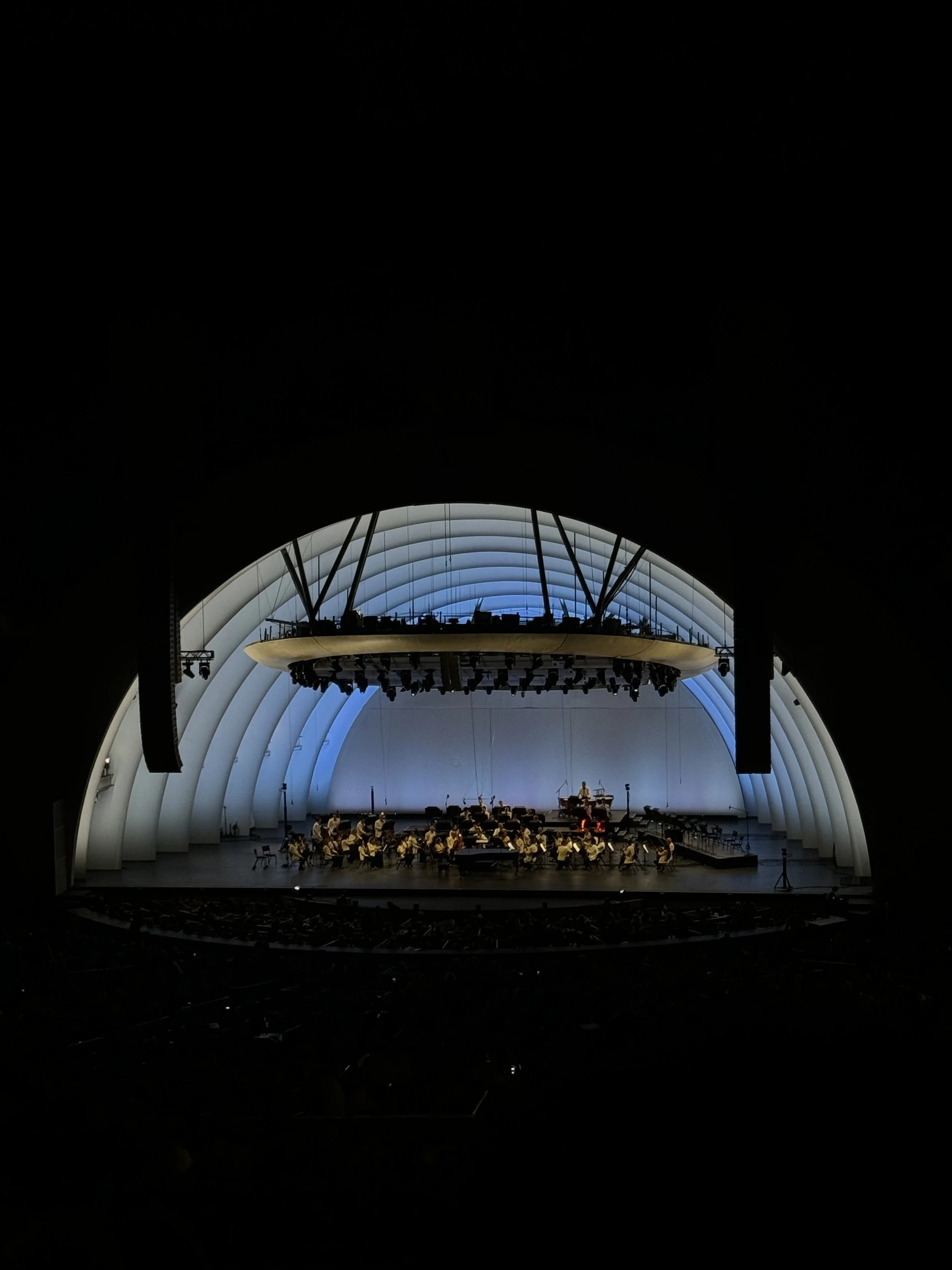 An indoor concert hall with a stage featuring an orchestra performing, illuminated with blue and white lighting, inside a curved, modern architectural structure.