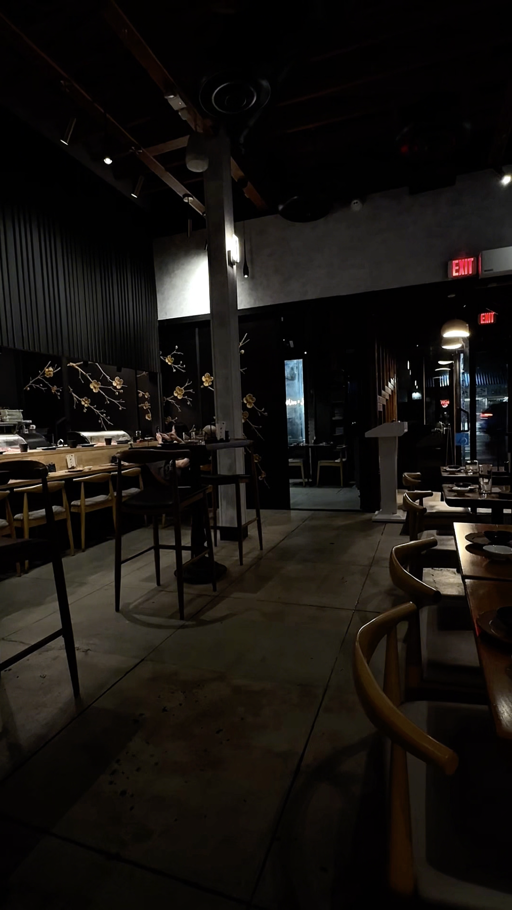 Interior of a dimly lit restaurant with wooden tables and chairs, decorative wall art of flowers, and illuminated exit signs near the entrance.