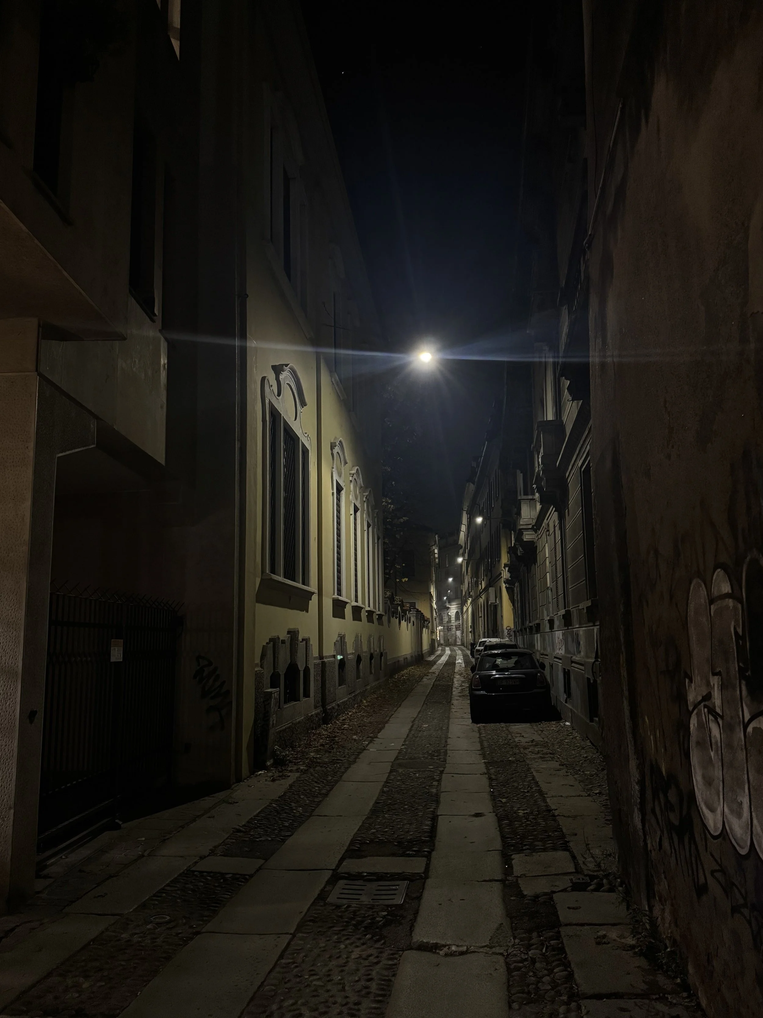 Nighttime view of a narrow street with parked cars, historic buildings on either side, and the moon visible in the dark sky.