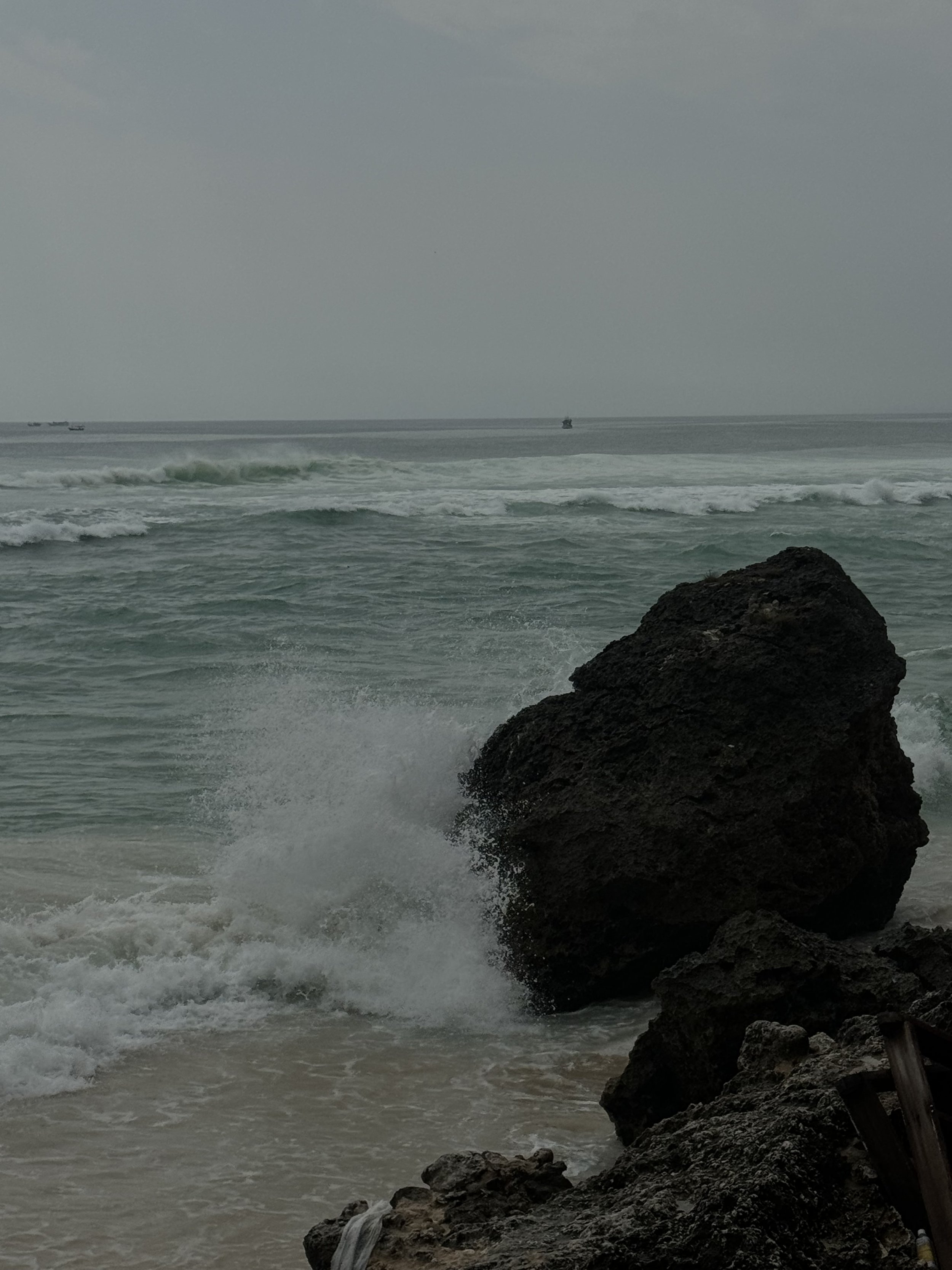 Overcast day at the ocean with waves crashing against rocks in the foreground; several boats visible in the distance on the water.