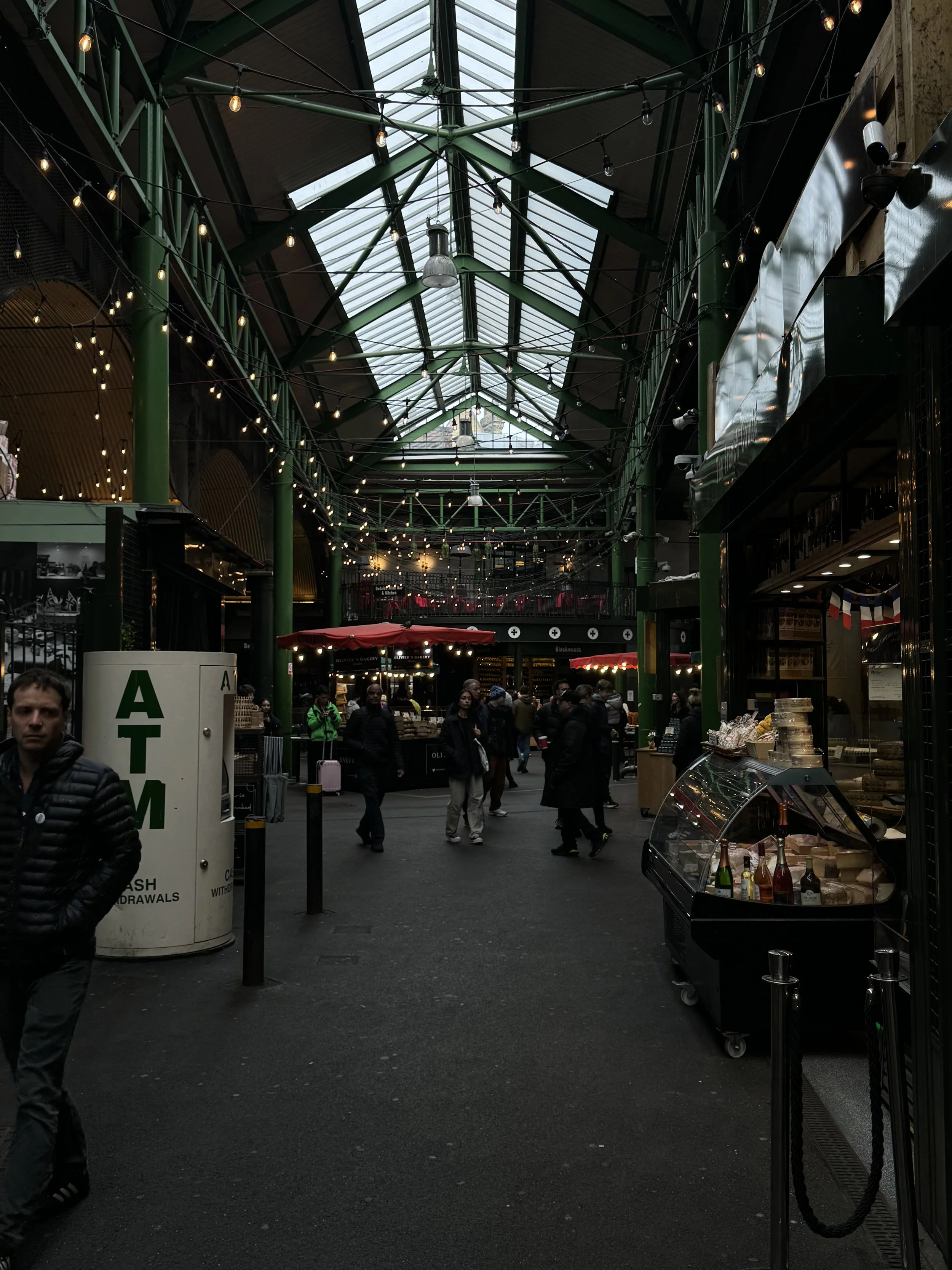 Indoor marketplace with high glass ceiling, green metal framework, string lights, and various food and retail stalls. Several people are walking and browsing.