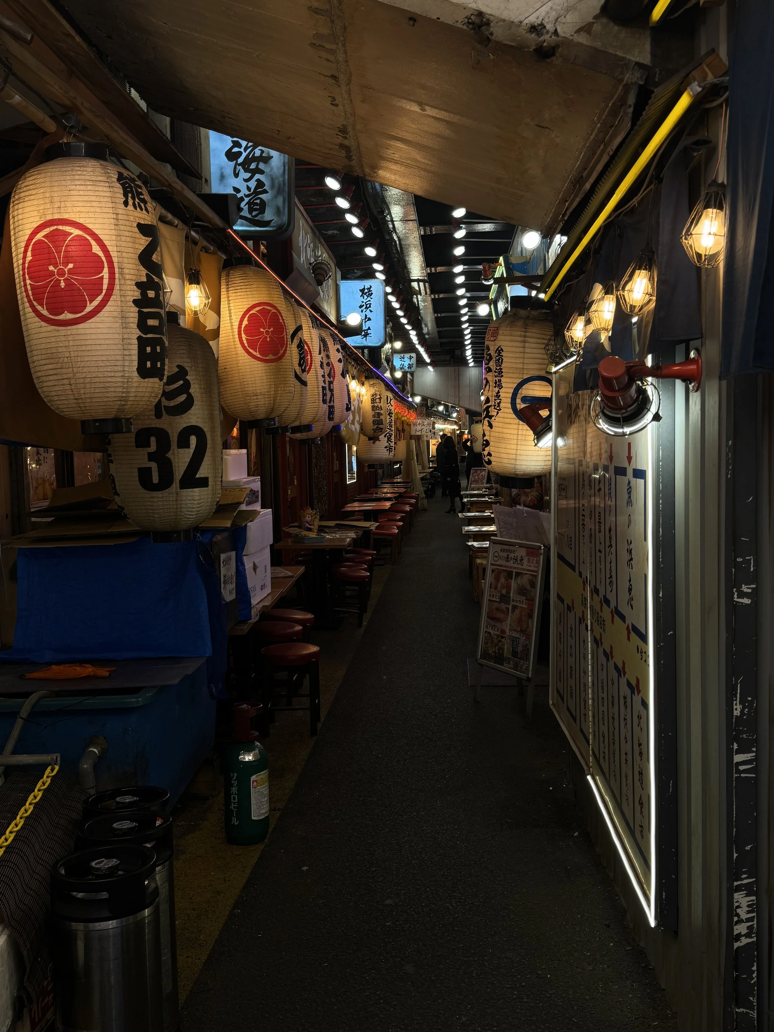 A narrow indoor corridor of a Japanese restaurant or izakaya, decorated with traditional paper lanterns, menu boards, and hanging signs, with wooden furniture and ambient lighting.