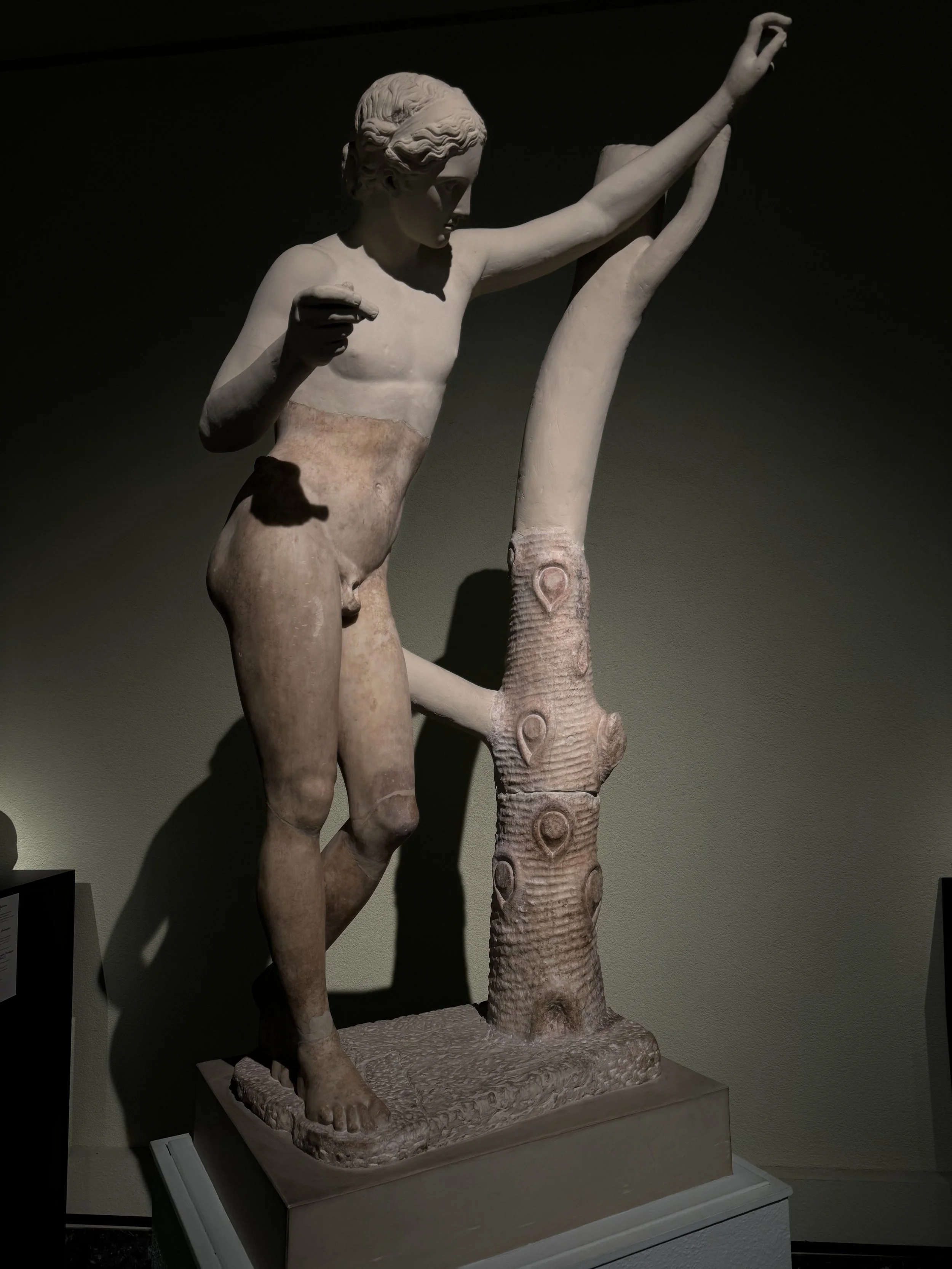 Ancient Greek sculpture of a standing male figure, partially draped, with one arm raised and holding a long staff with a carved pattern, displayed on a pedestal in a museum setting.