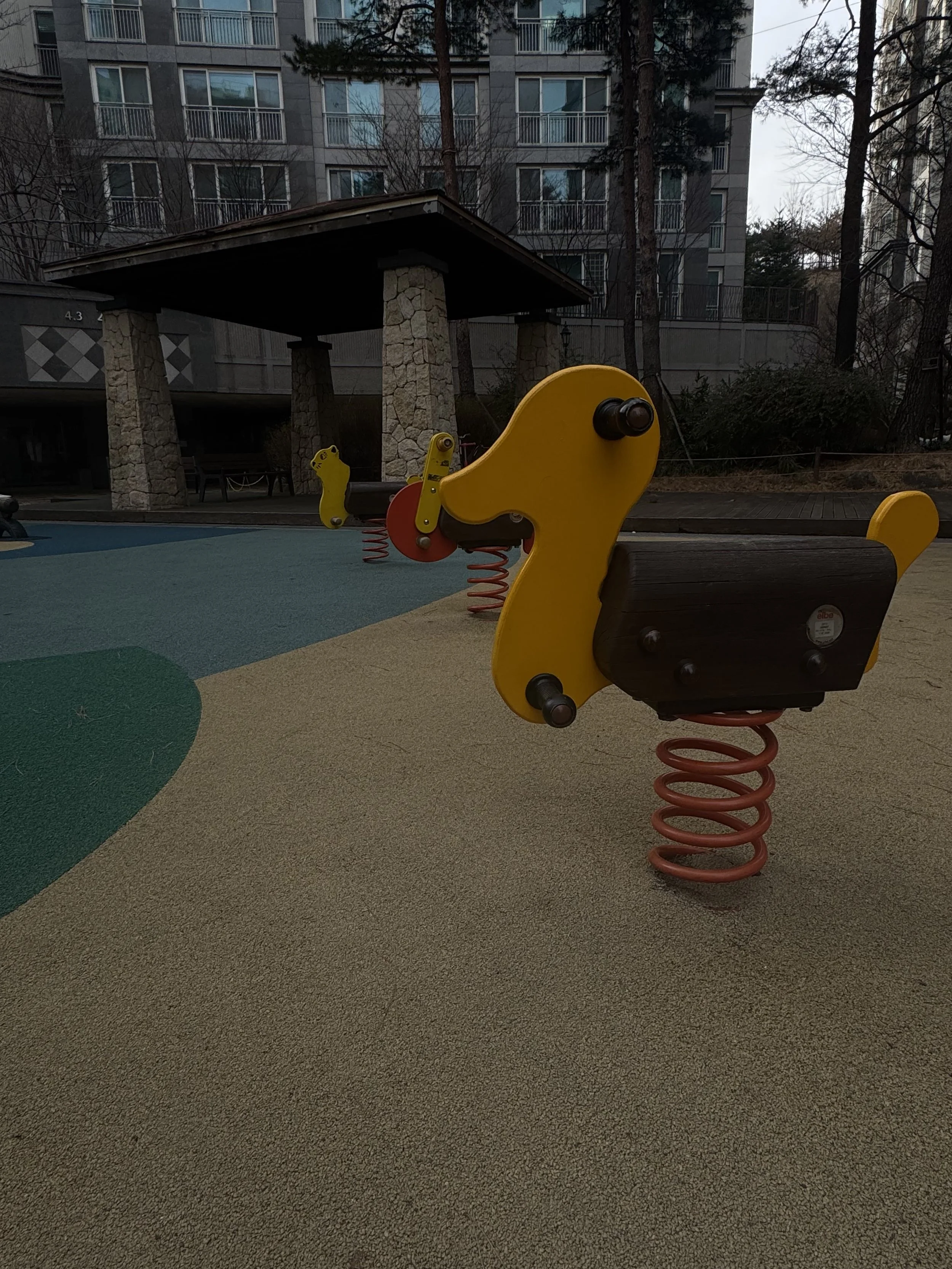 Empty playground with a yellow animal-shaped spring rocker in the foreground, a stone shelter with a roof, benches, and a multi-story residential building with balconies in the background.