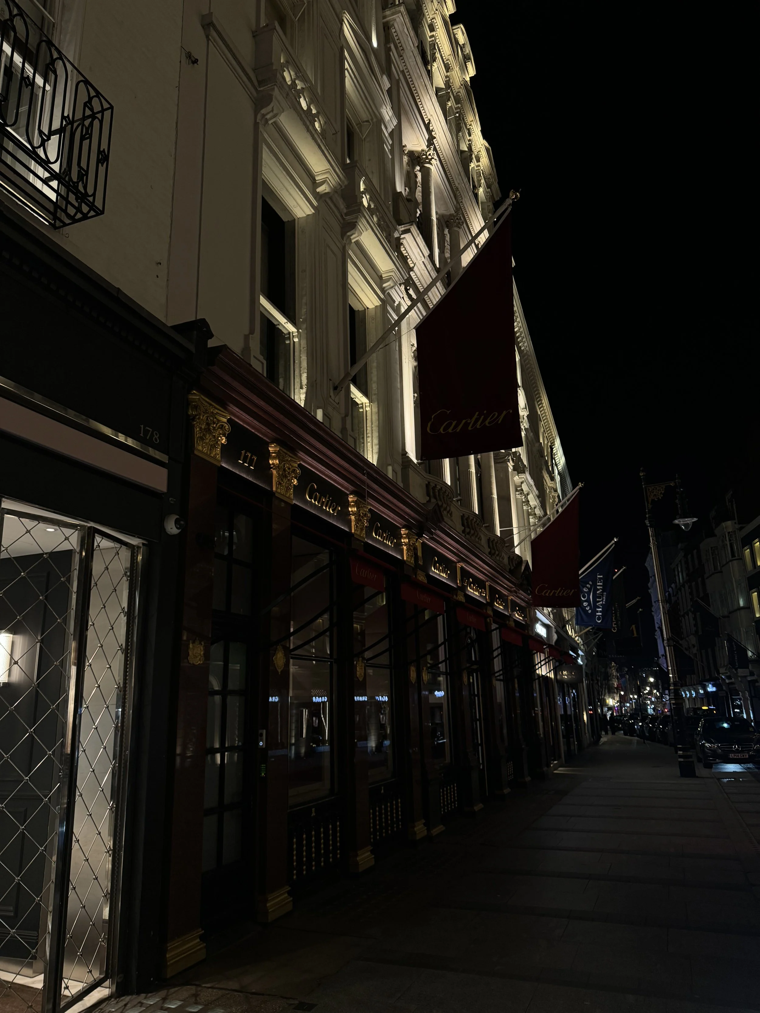 Night view of a luxury storefront, Cartier, with ornate architecture and flags outside, illuminated in the dark with city street lights in the background.