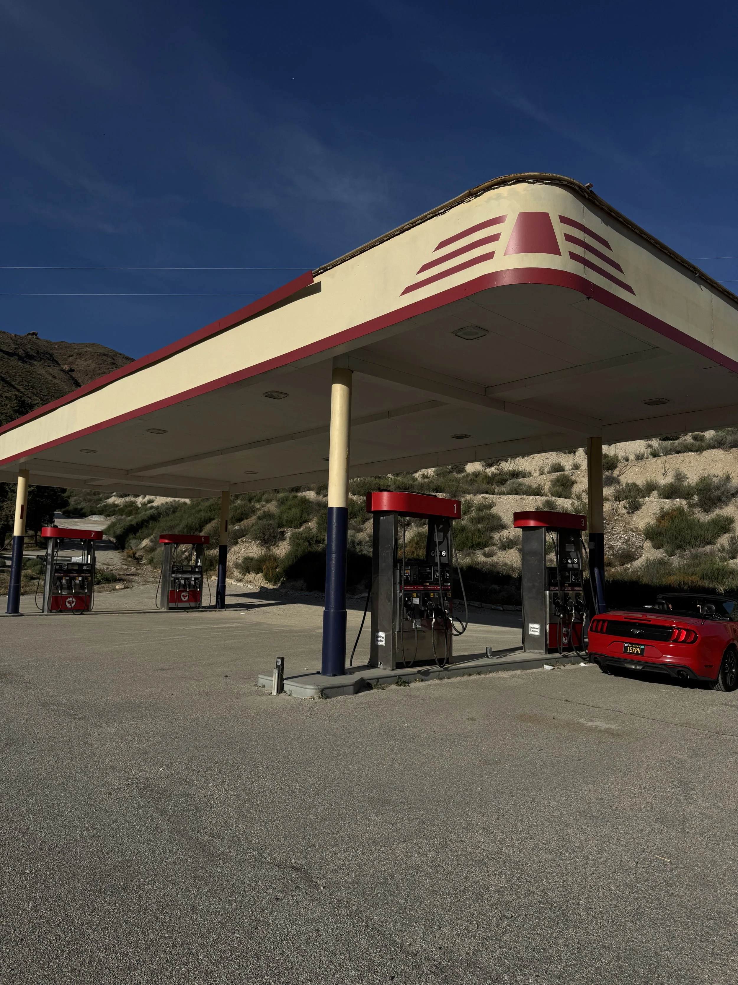 Empty gas station with four fuel pumps, a red sports car parked nearby, and mountainous terrain in the background under a clear blue sky.