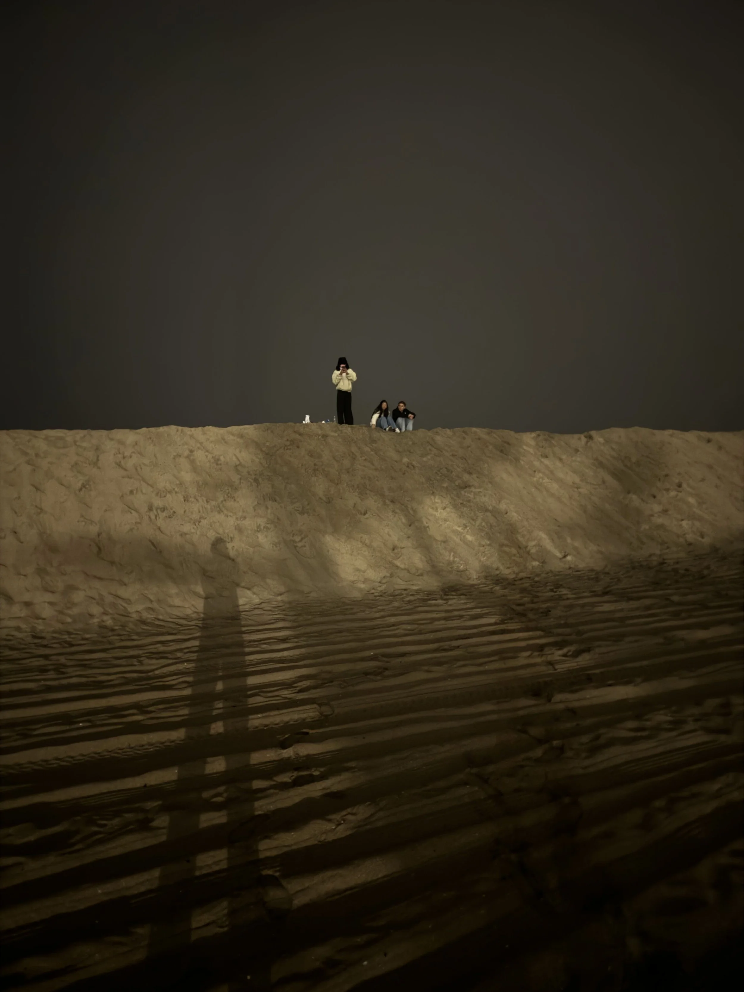 Three people standing and sitting on a sandy beach at night, with a dark sky above and a wooden pier in the foreground.