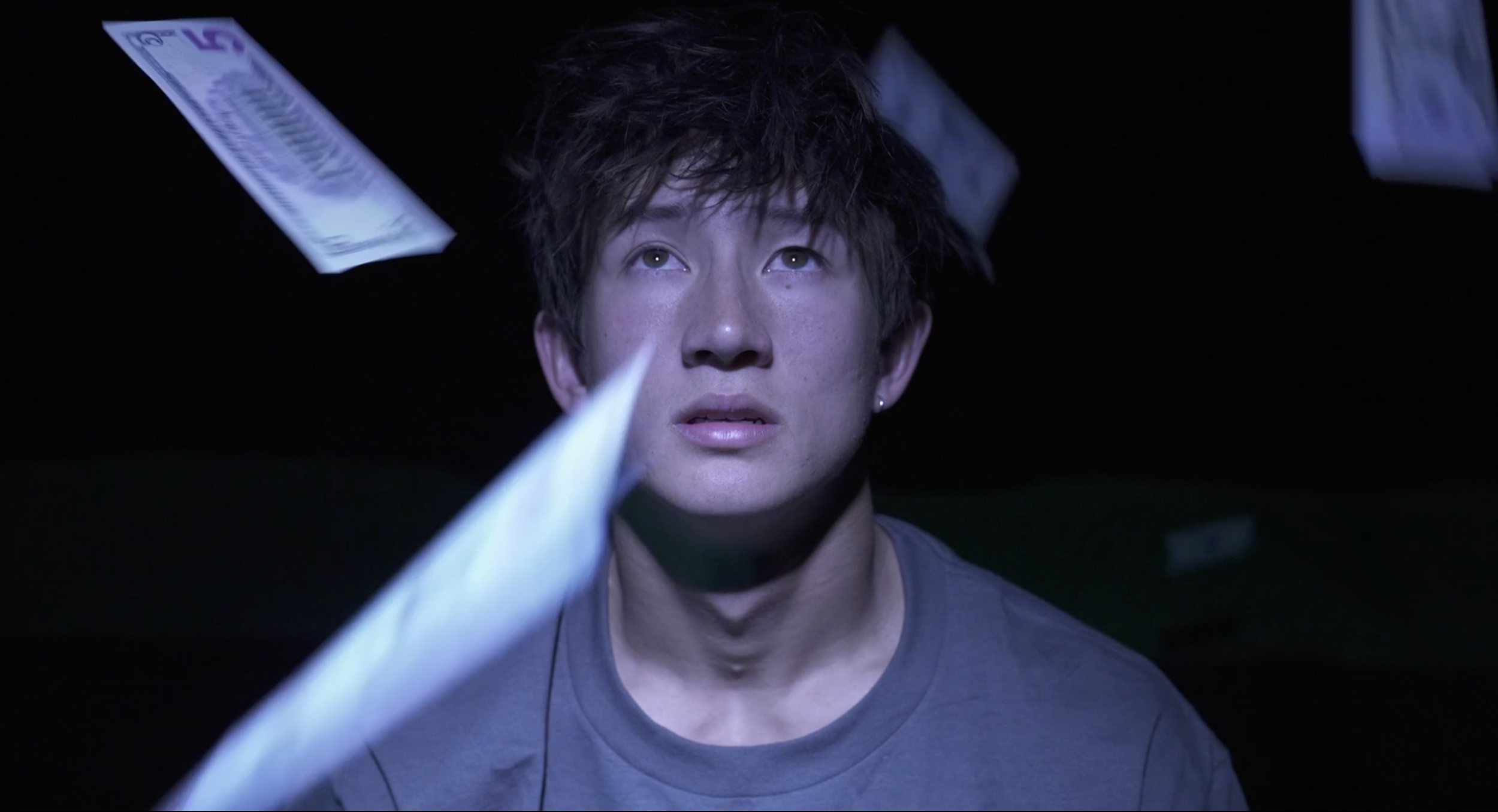 A young person with short dark hair and earrings, looking upward with a surprised or curious expression, surrounded by floating white and purple paper or dollar bills at night or in a dark setting.