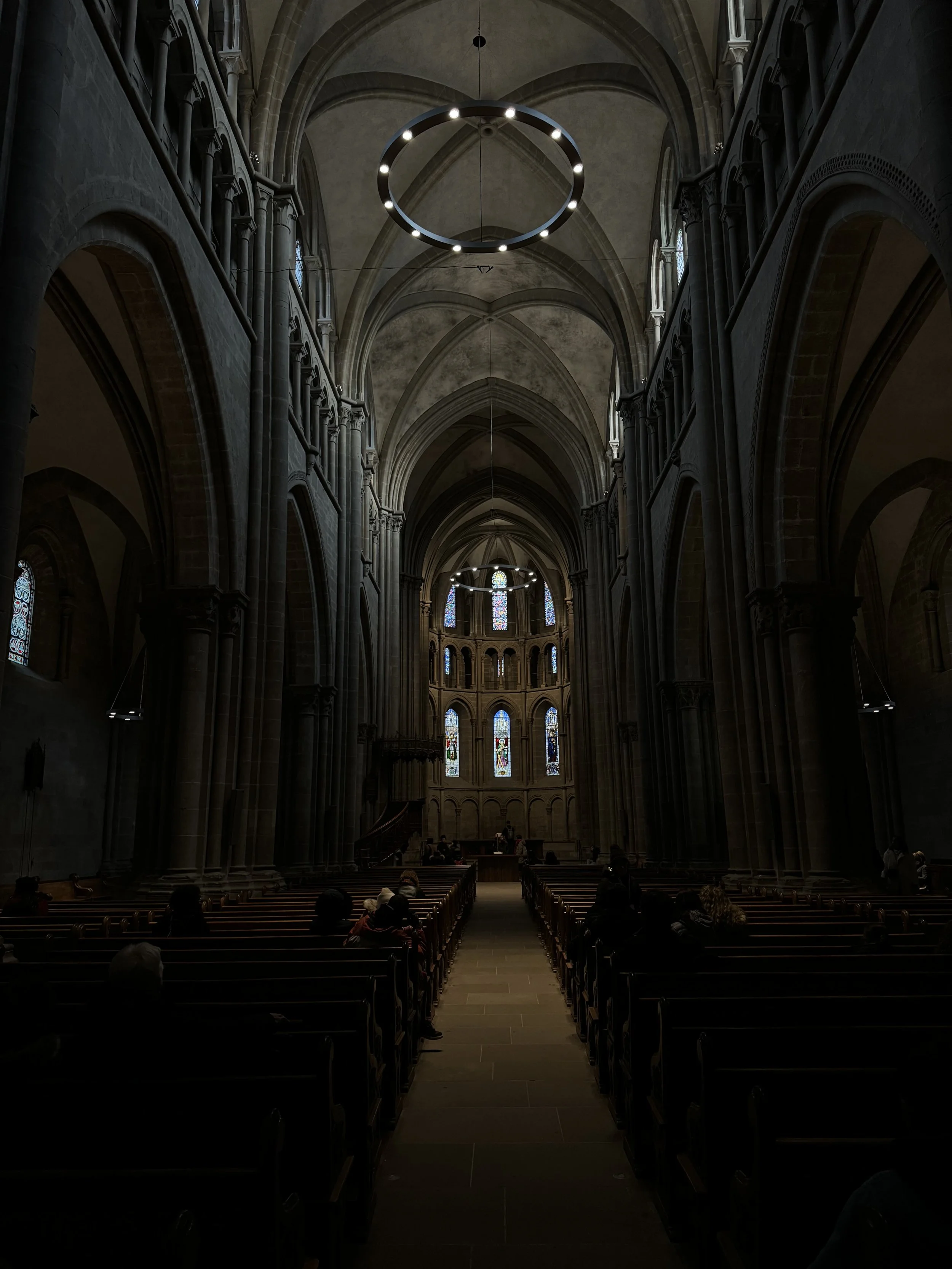 Interior of a Gothic-style church with high vaulted ceilings, large arched windows with stained glass, and hanging circular chandeliers, with people sitting on wooden pews.