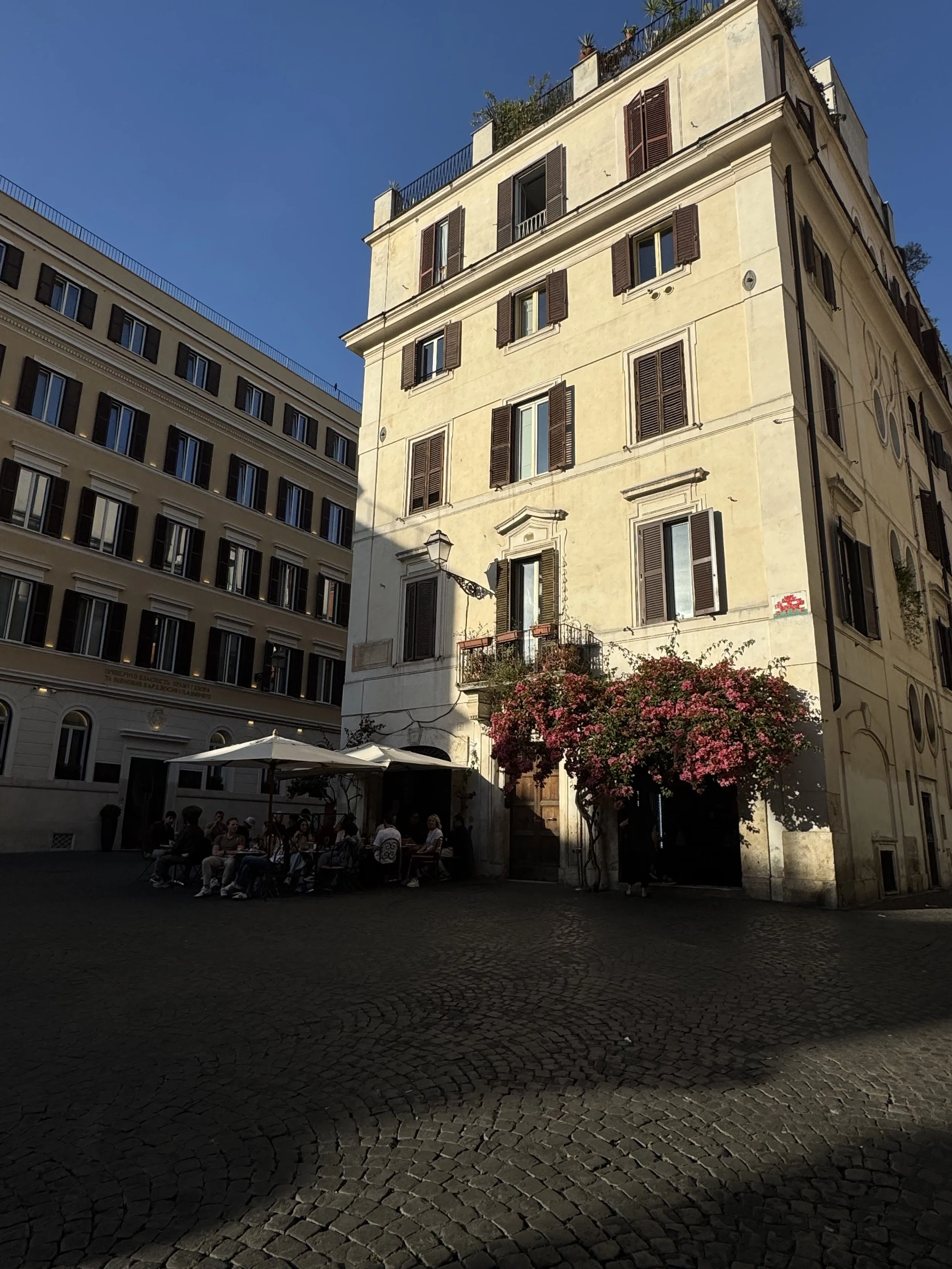 A group of people sitting at an outdoor cafe under an umbrella in a European city square, with old buildings and a cobblestone street, contrasted by bright sunlight and a blue sky.
