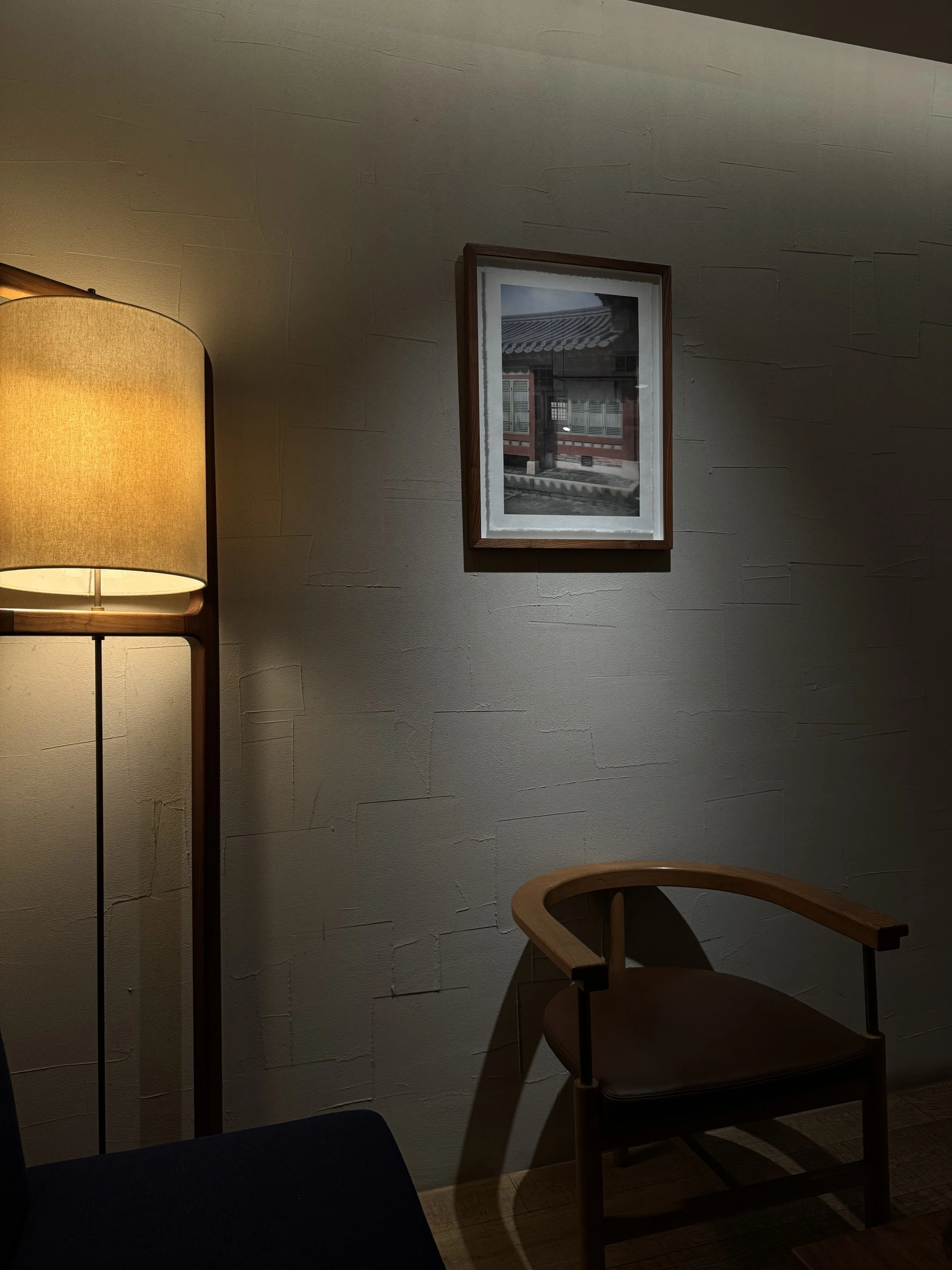 Interior scene with a wooden chair, a floor lamp with a yellow lampshade, and a framed picture of a traditional Asian building on a textured beige wall.