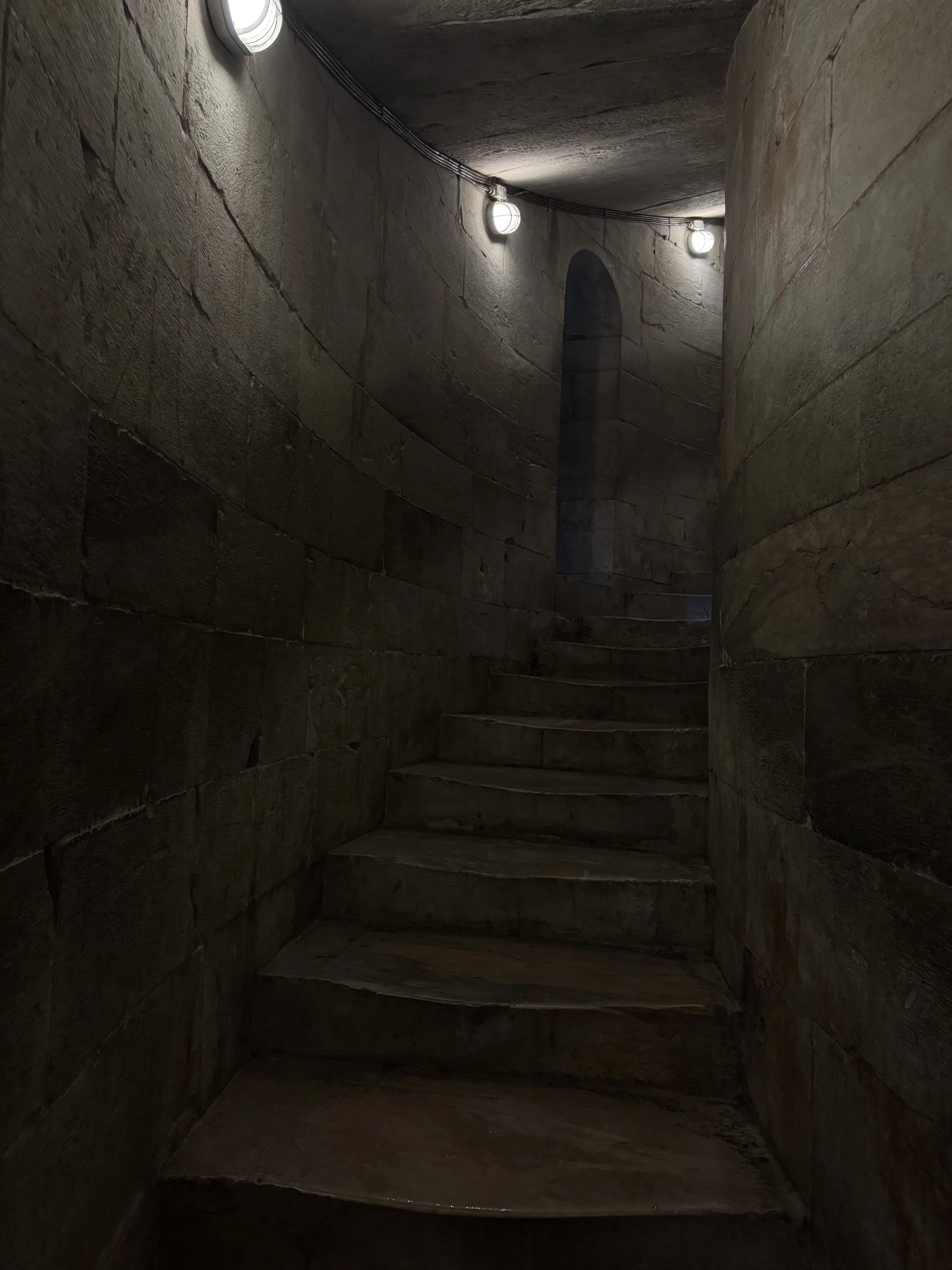 A narrow stone staircase inside a medieval stone tower with arched window at the top, illuminated by wall-mounted lights.
