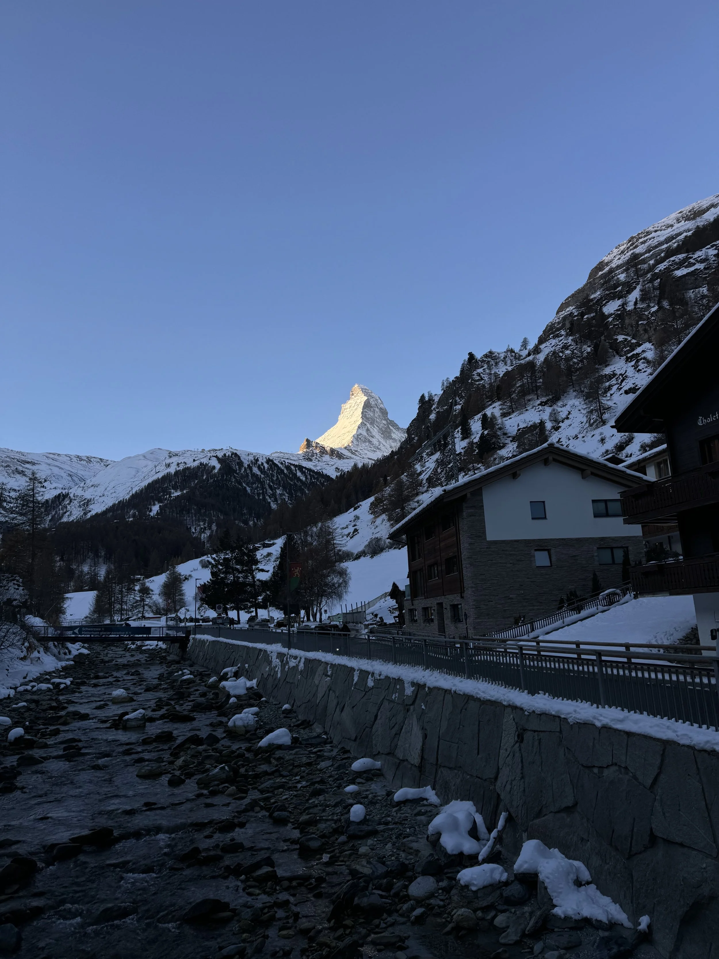 Snow-covered mountains with a prominent peak in the background, a small village with buildings, a road, and a stream with snow on its banks in the foreground, and a clear blue sky.