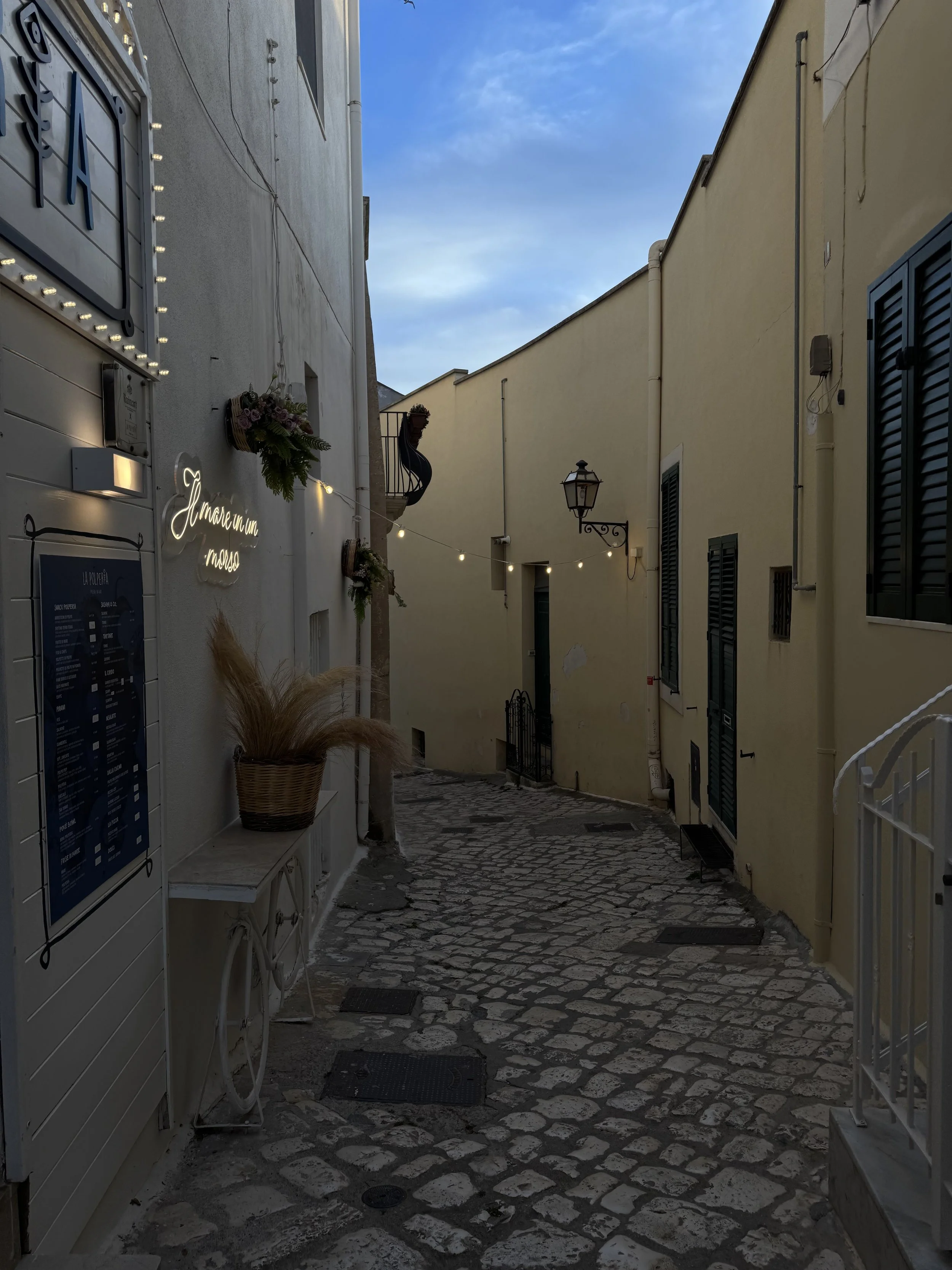 A narrow cobblestone street in a European town with pastel-colored buildings on either side. There is a flower basket, string lights, and a neon sign on the left wall, and a street lamp on the right.