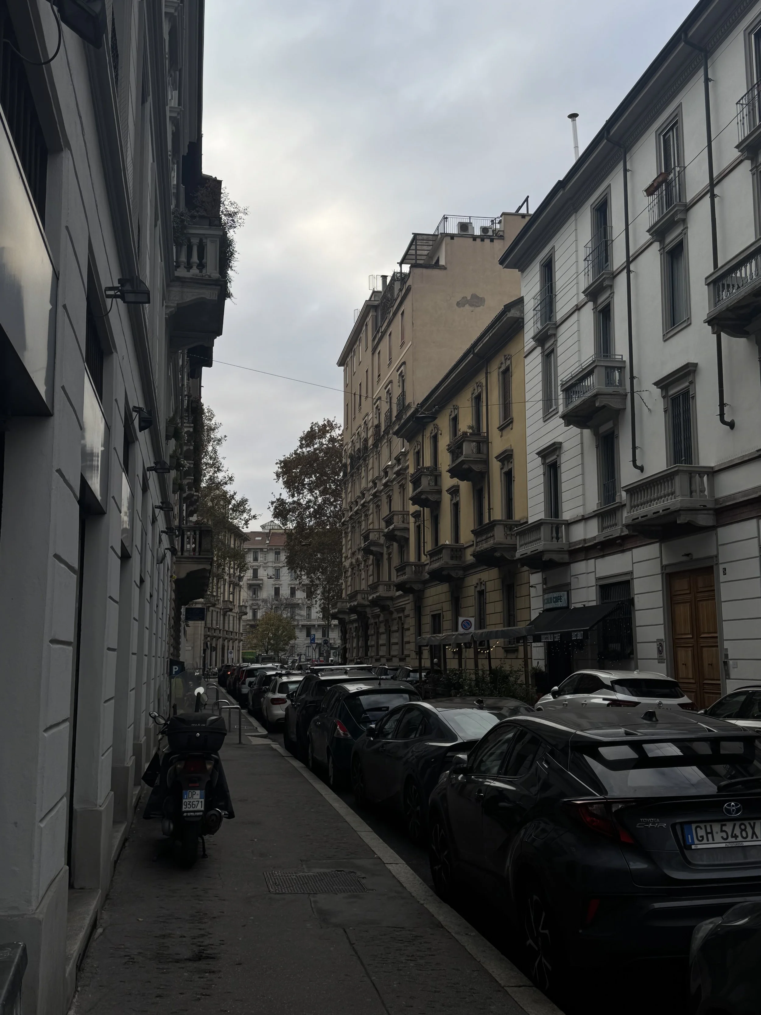 View of a city street with parked cars along the side, multi-story buildings with balconies, and a cloudy sky overhead.