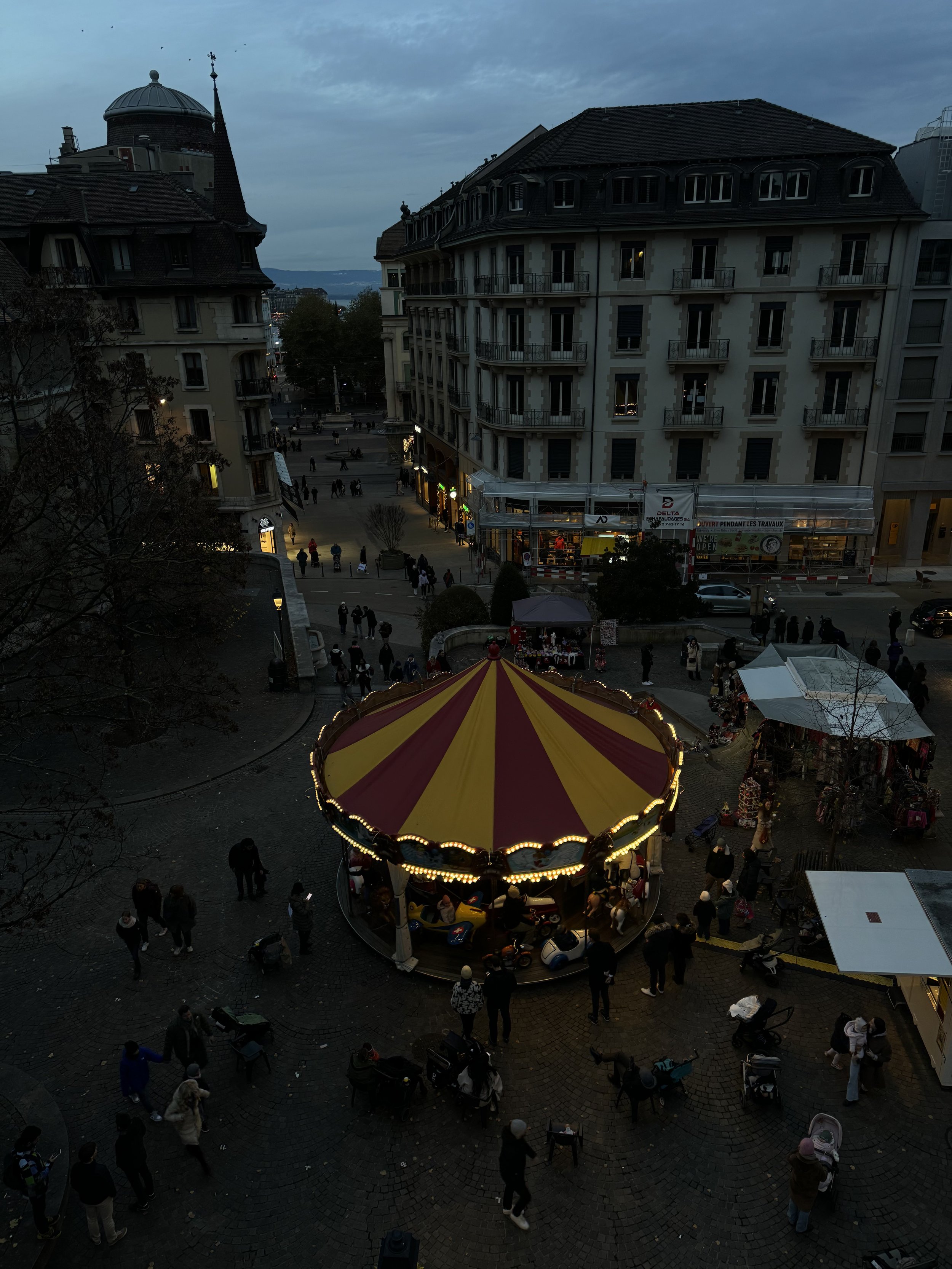 A carousel with a red and yellow striped canopy in a busy town square during the evening. People walk around, some with strollers. Surrounding buildings include shops and apartment complexes, with a cloudy sky overhead.