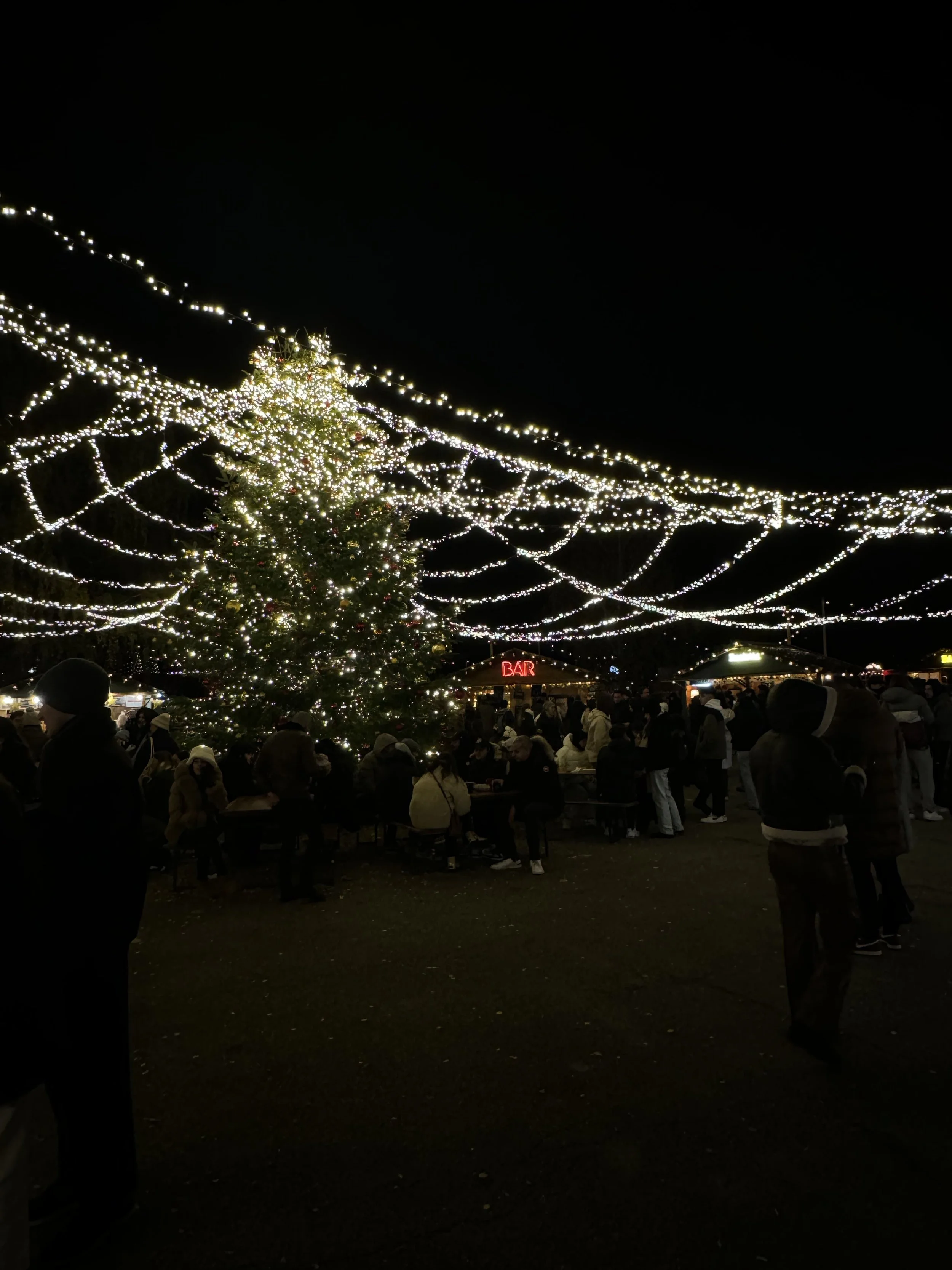 A large decorated Christmas tree surrounded by people at an outdoor night event, with string lights overhead and a bar in the background.