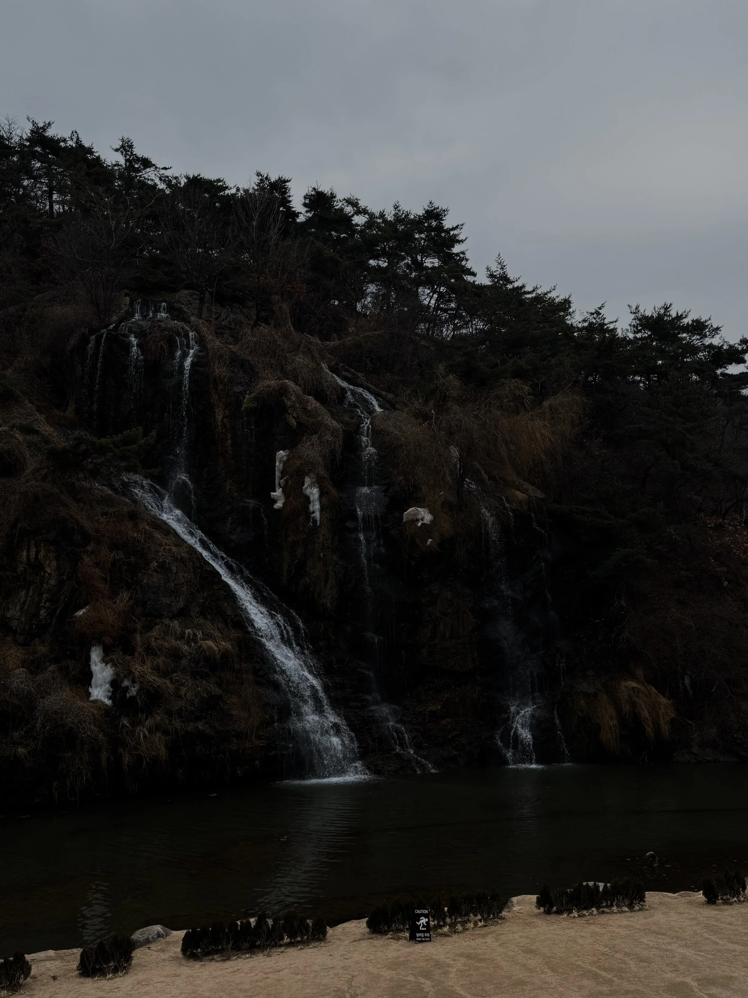 A waterfall cascading down a rocky hillside with trees on top, over a body of water, and a small beach area in the foreground with bushes and a caution sign.