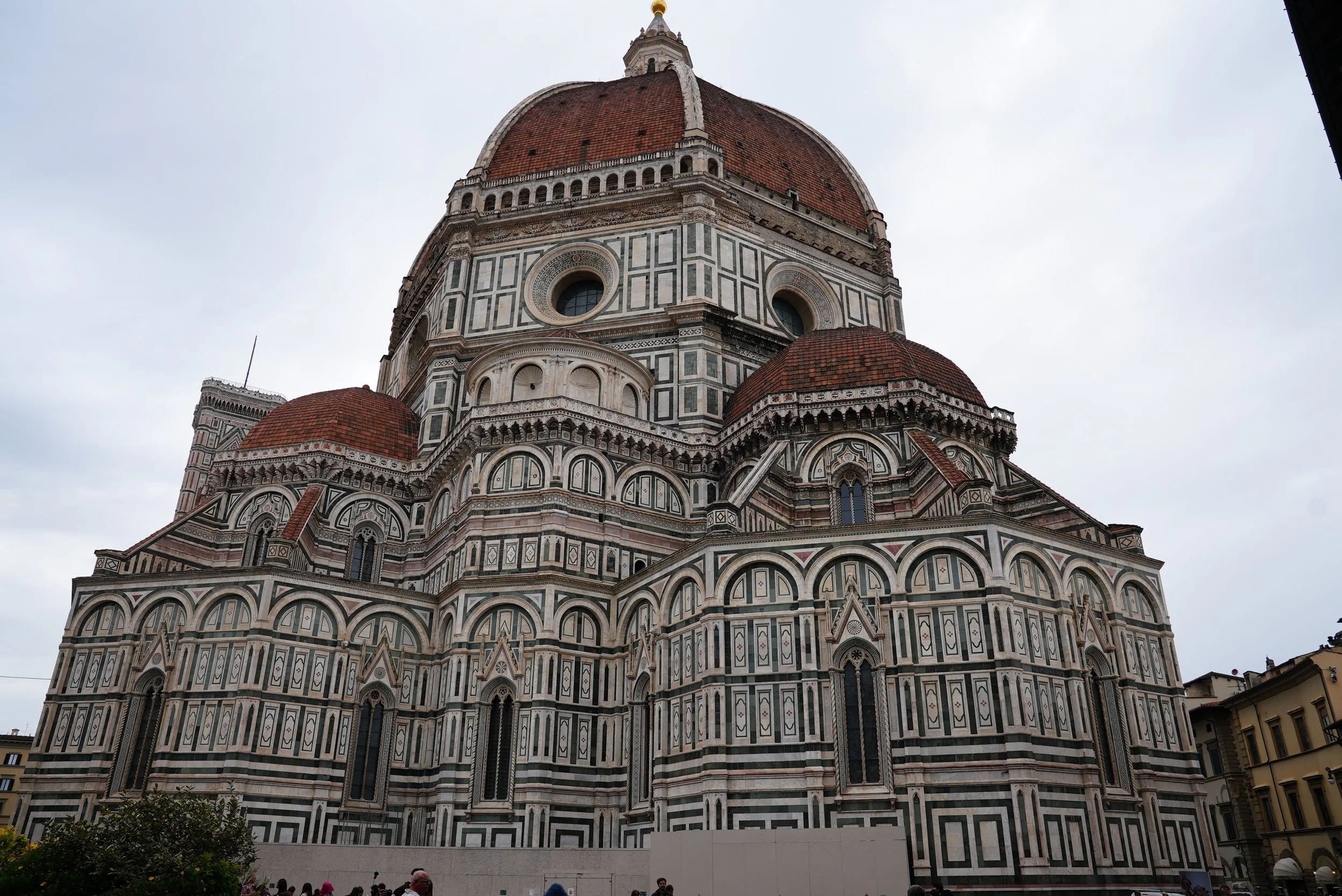 A large, ornate cathedral with a distinctive green, white, and pink marble patterned exterior. The cathedral has a large dome with a red-tiled roof and smaller domes surrounding it. There are windows and decorative arches on the facade, and a crowd o