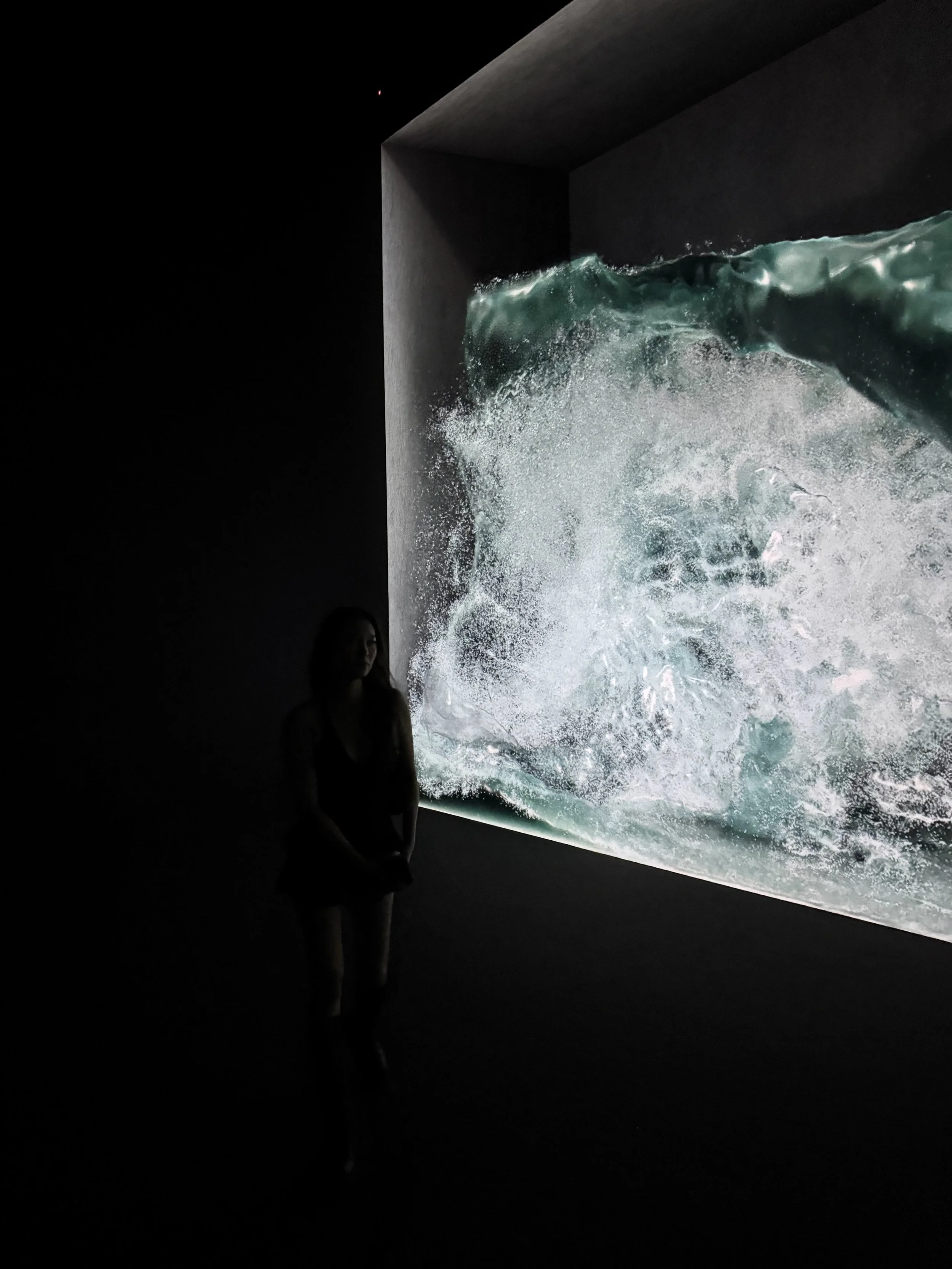 Dark room with a large screen displaying a detailed image of an ocean wave, with a person standing in front of it.