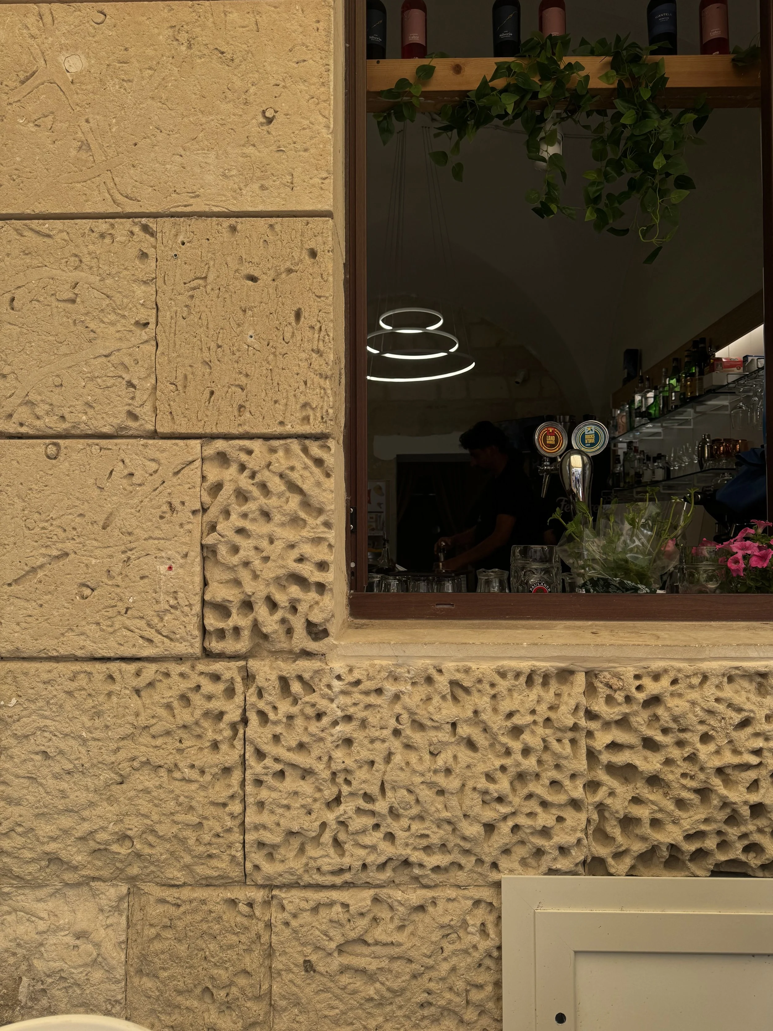 The image shows a close-up view of a textured beige stone wall and a window with a wooden frame. Inside the window, a person with dark hair is visible, working behind a bar or counter. There are bottles on shelves and modern circular ceiling lights h