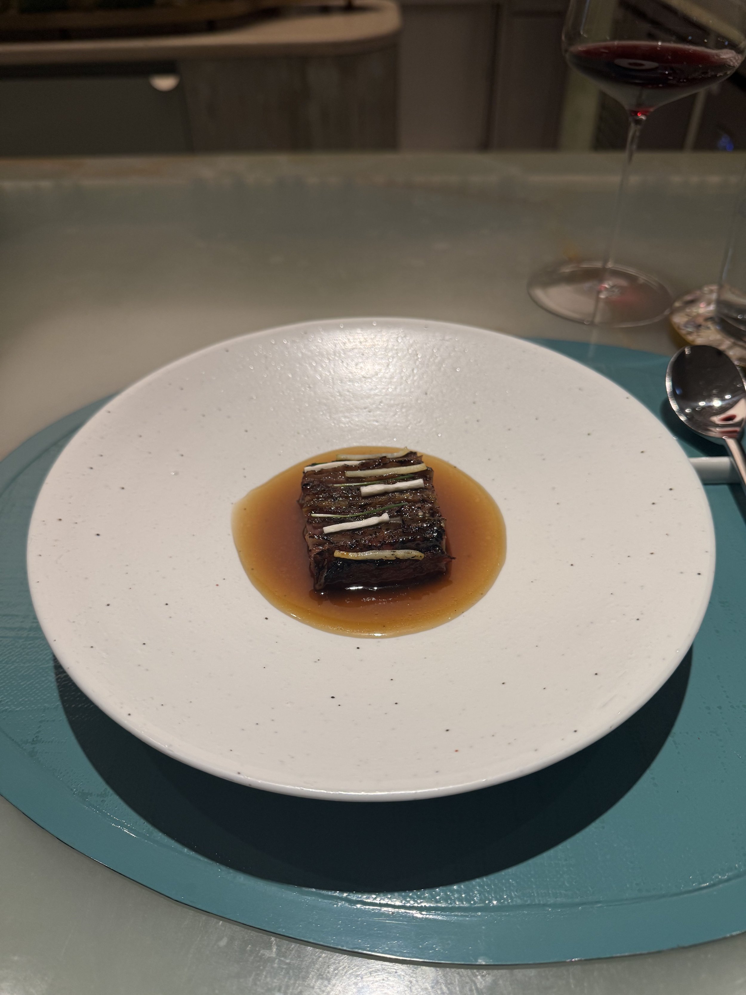 A plated dish with a piece of grilled meat topped with thinly sliced green onions, served with a brown sauce on a white plate. Two glasses of red wine are visible in the background.
