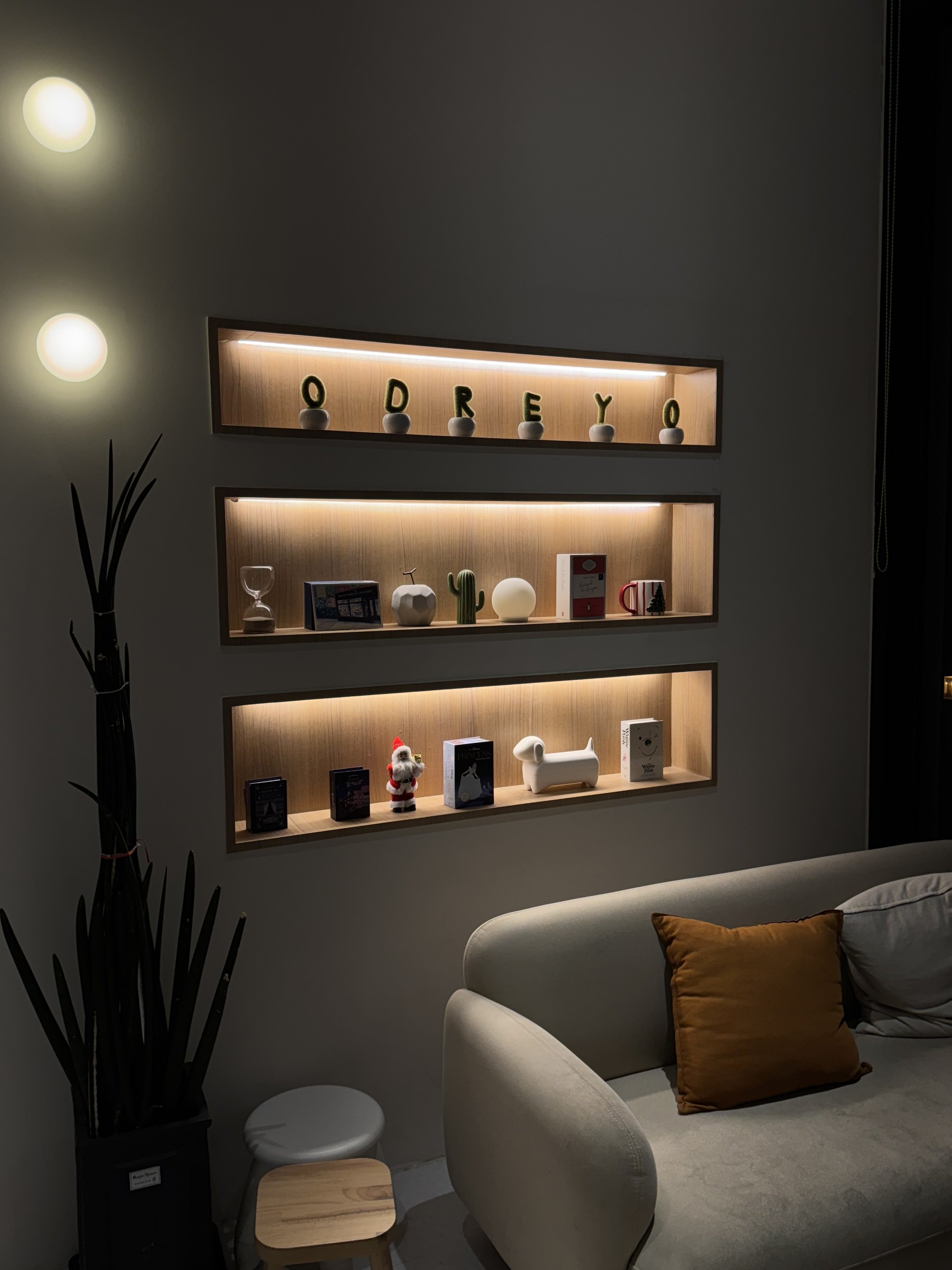 Decorative wall with three illuminated recessed shelves displaying small decorative objects and books, adjacent to a white sofa with orange and gray cushions, and a floor lamp with a black stand and abstract design.