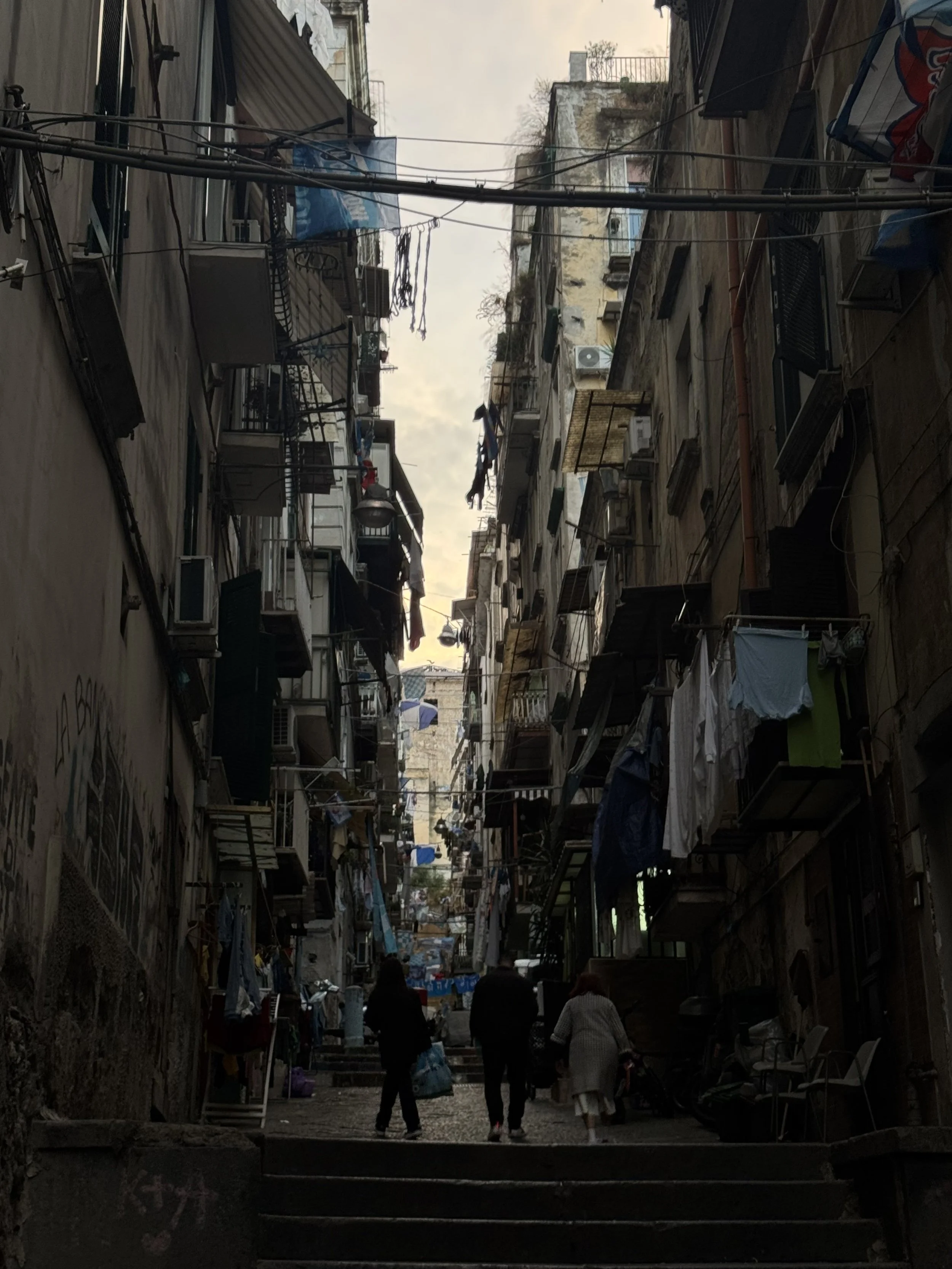 A narrow urban street scene with several people walking, surrounded by apartment buildings with laundry hanging on lines, and electrical wires overhead.