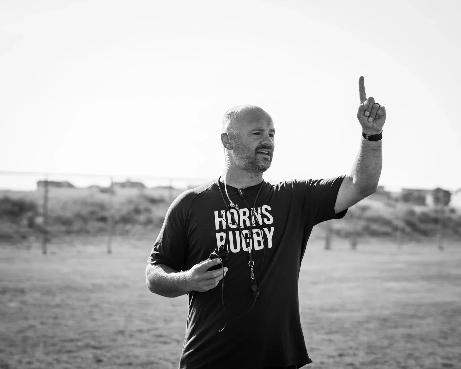 A man with a beard and short hair, wearing a black t-shirt with the slogan 'HORNS RUGBY', is standing outdoors on a field with a blurred background of houses and trees. He is pointing upward with his right hand and holding a stopwatch or timer in his left hand, with a whistle around his neck.