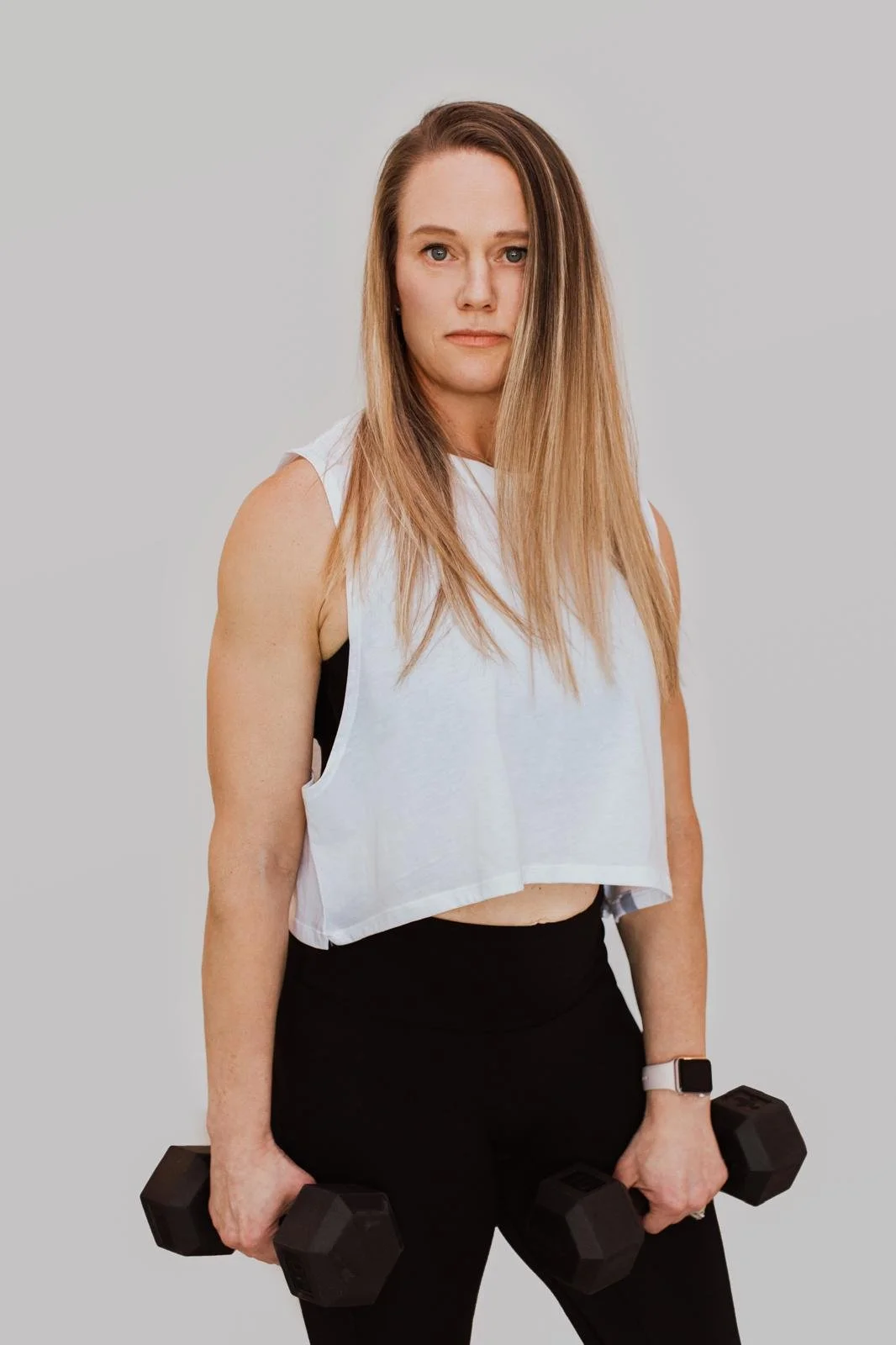 A woman with long blonde hair wearing a sleeveless white sports top, black leggings, and a smartwatch, holding black dumbbells in both hands, standing against a plain light grey background.
