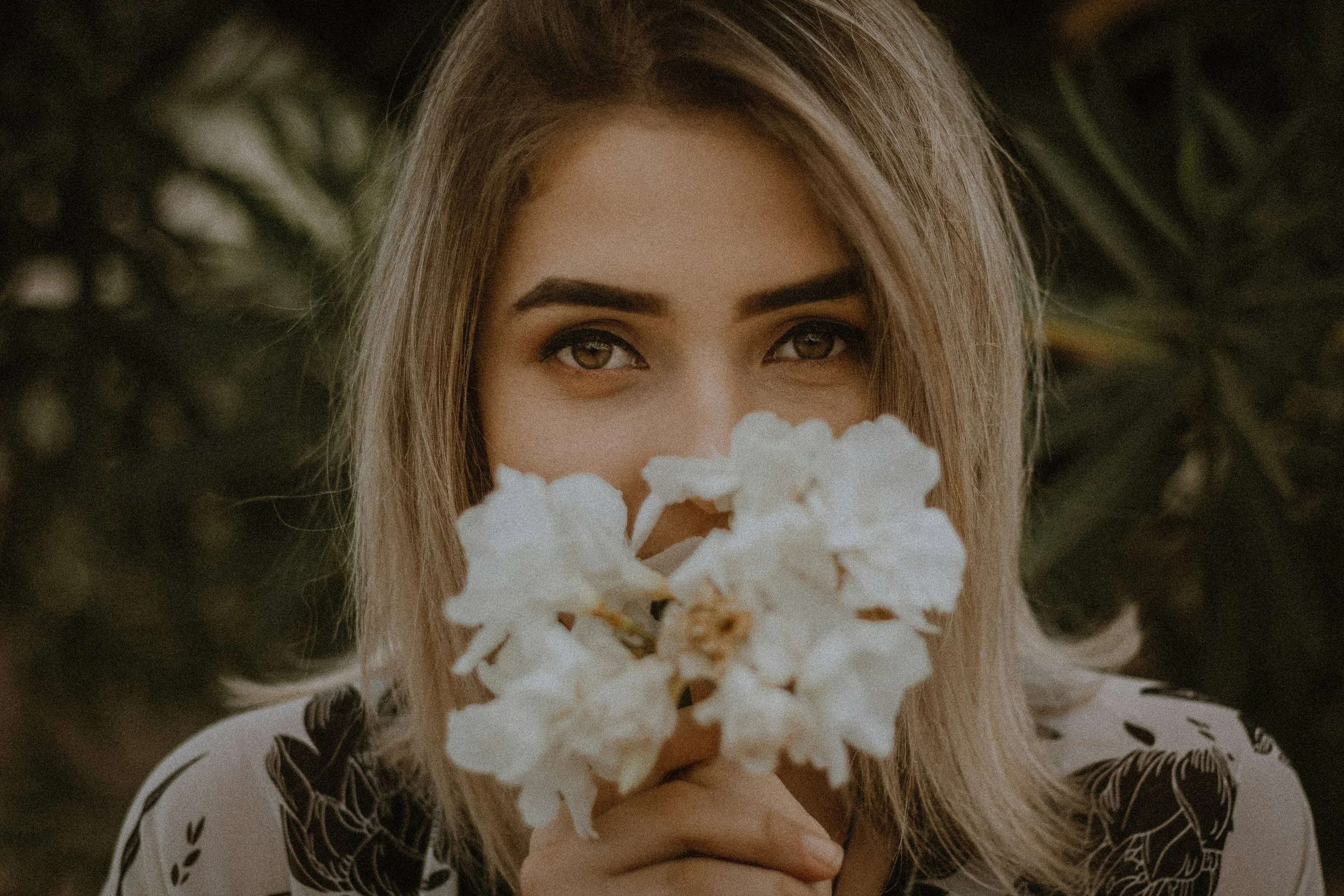 Close-up portrait of a blonde woman holding white flowers in front of her face outdoors